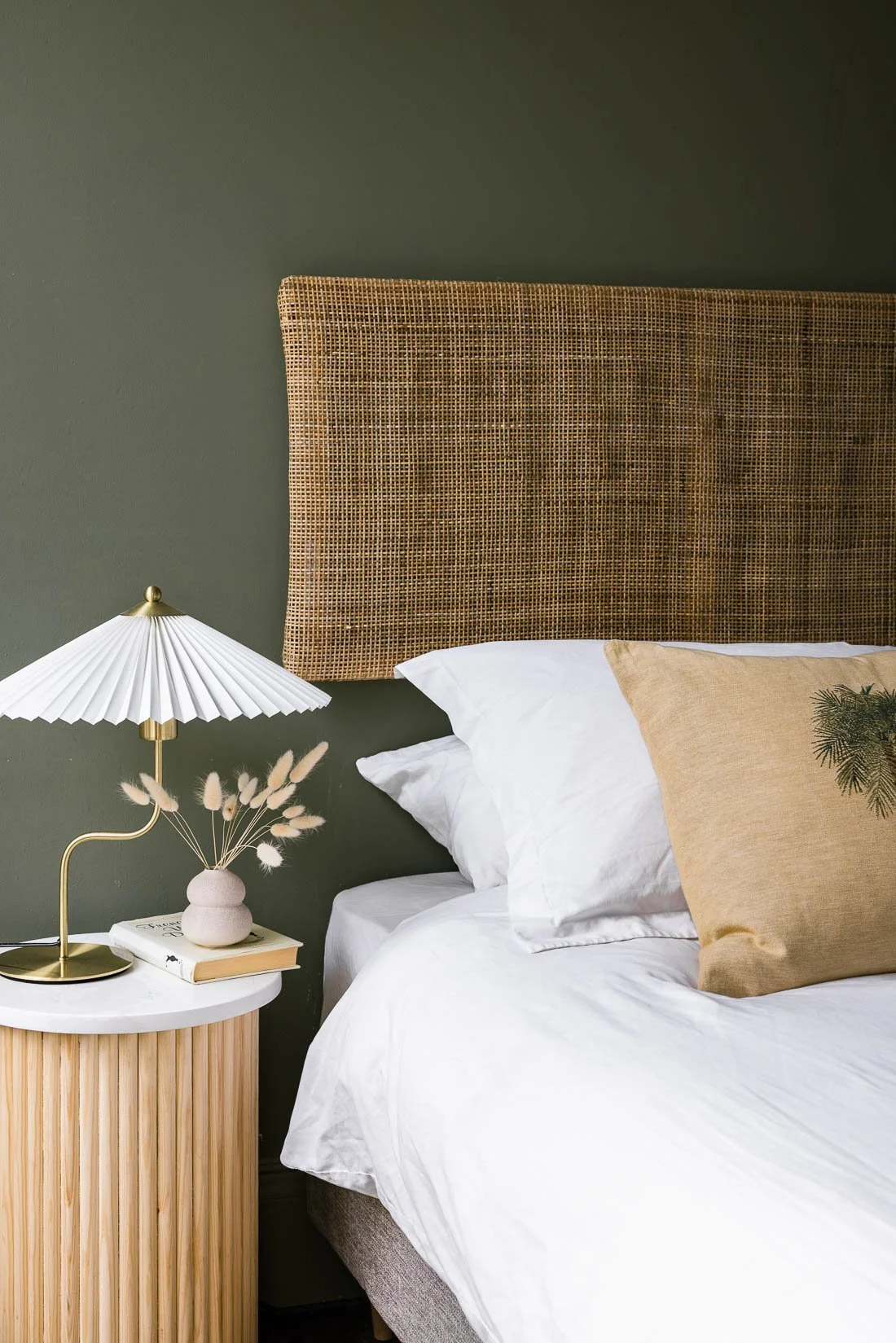 Bedroom with a green wall, a rattan headboard, a white bed with white and beige pillows, a round white bedside table with a white and gold lamp, a small ceramic vase with dried flowers, and a book.