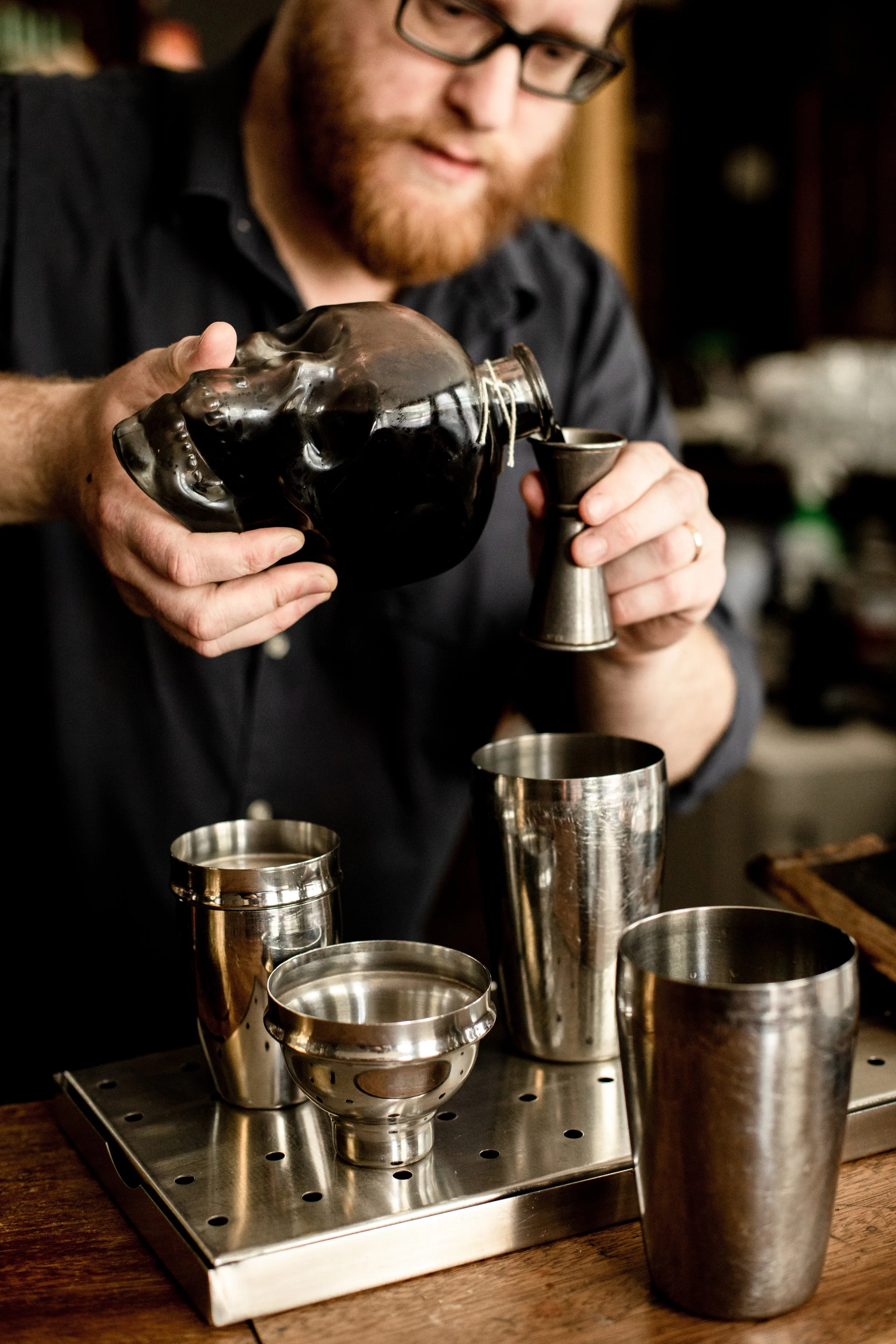 A man with glasses pouring a dark beverage from a glass skull-shaped bottle into a metal jigger, with various metal cocktail shakers and a perforated metal strainer on a bar counter.