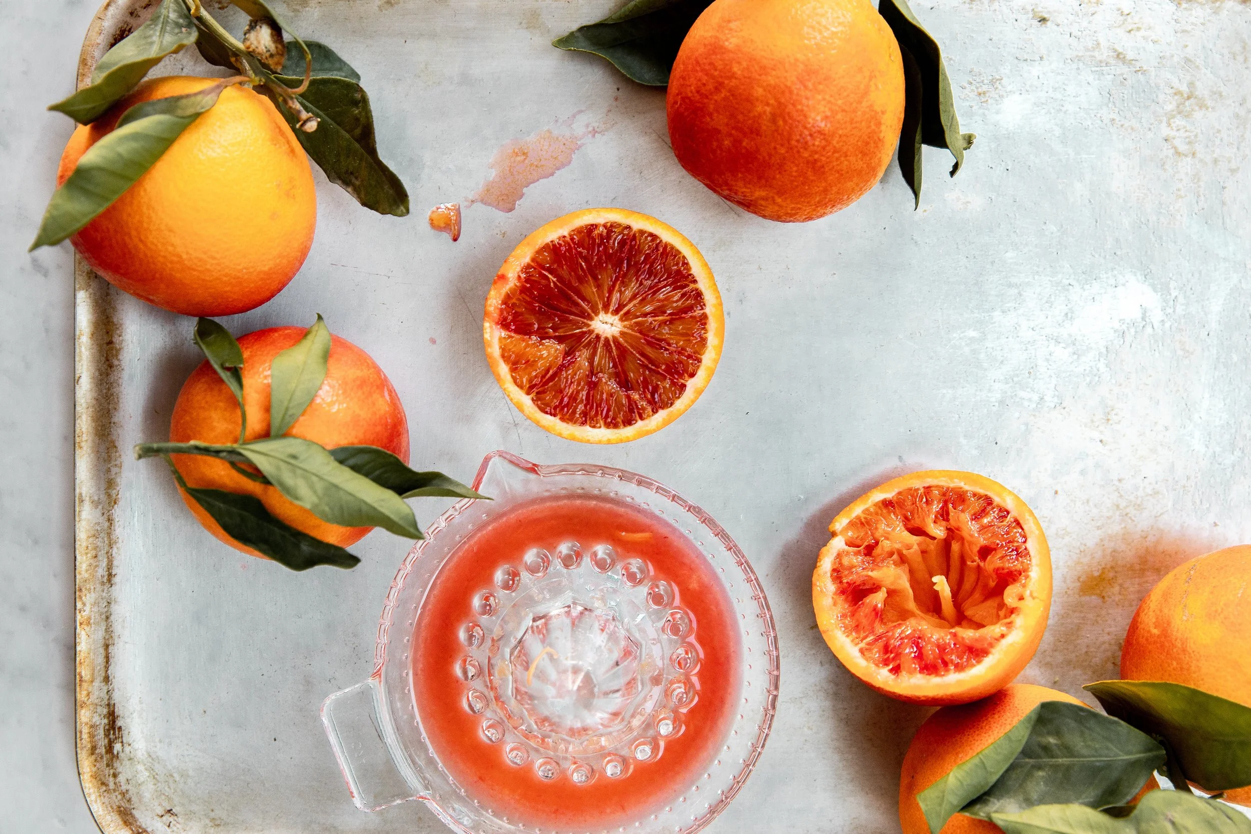 Several whole oranges with green leaves, one partially peeled orange, and a halved orange showing its juicy interior, arranged on a light-colored surface with a glass of pink grapefruit juice in the foreground.
