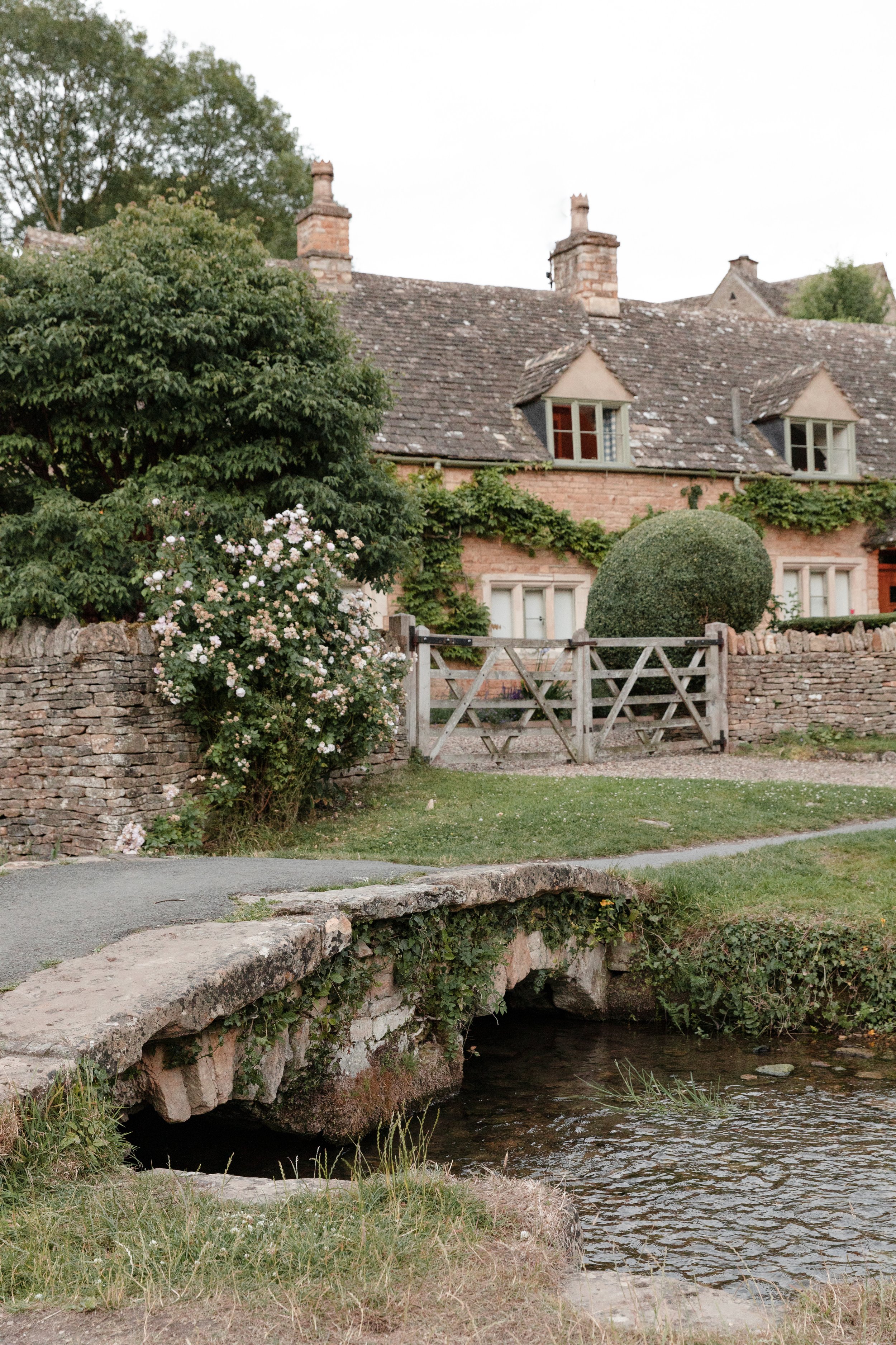 A stone bridge over a small creek, with a brick house featuring a tiled roof, dormer windows, and chimney stacks in the background, surrounded by green trees and bushes.