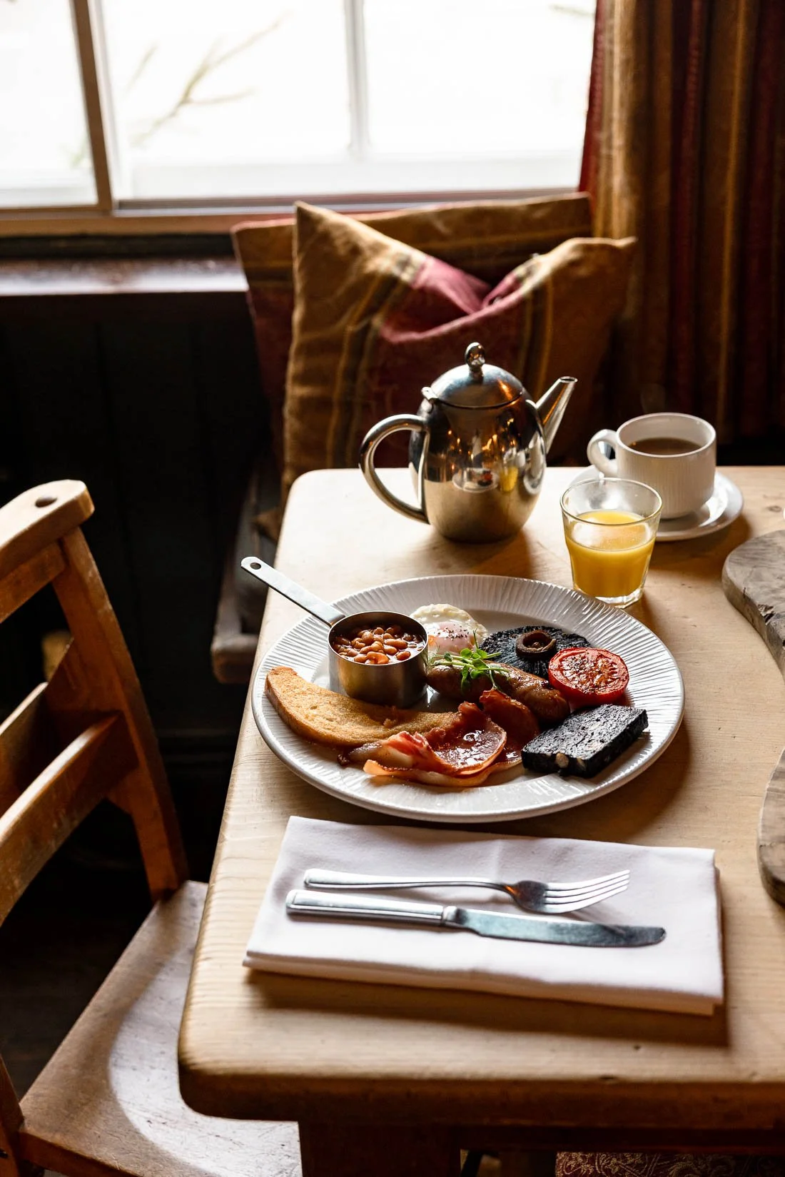 A breakfast spread on a wooden table includes a plate with baked beans, toast, scrambled eggs, bacon, tomatoes, black pudding, and several meats. There is a small glass of orange juice, a cup of coffee, a teapot, and a cup with saucer. A napkin with 