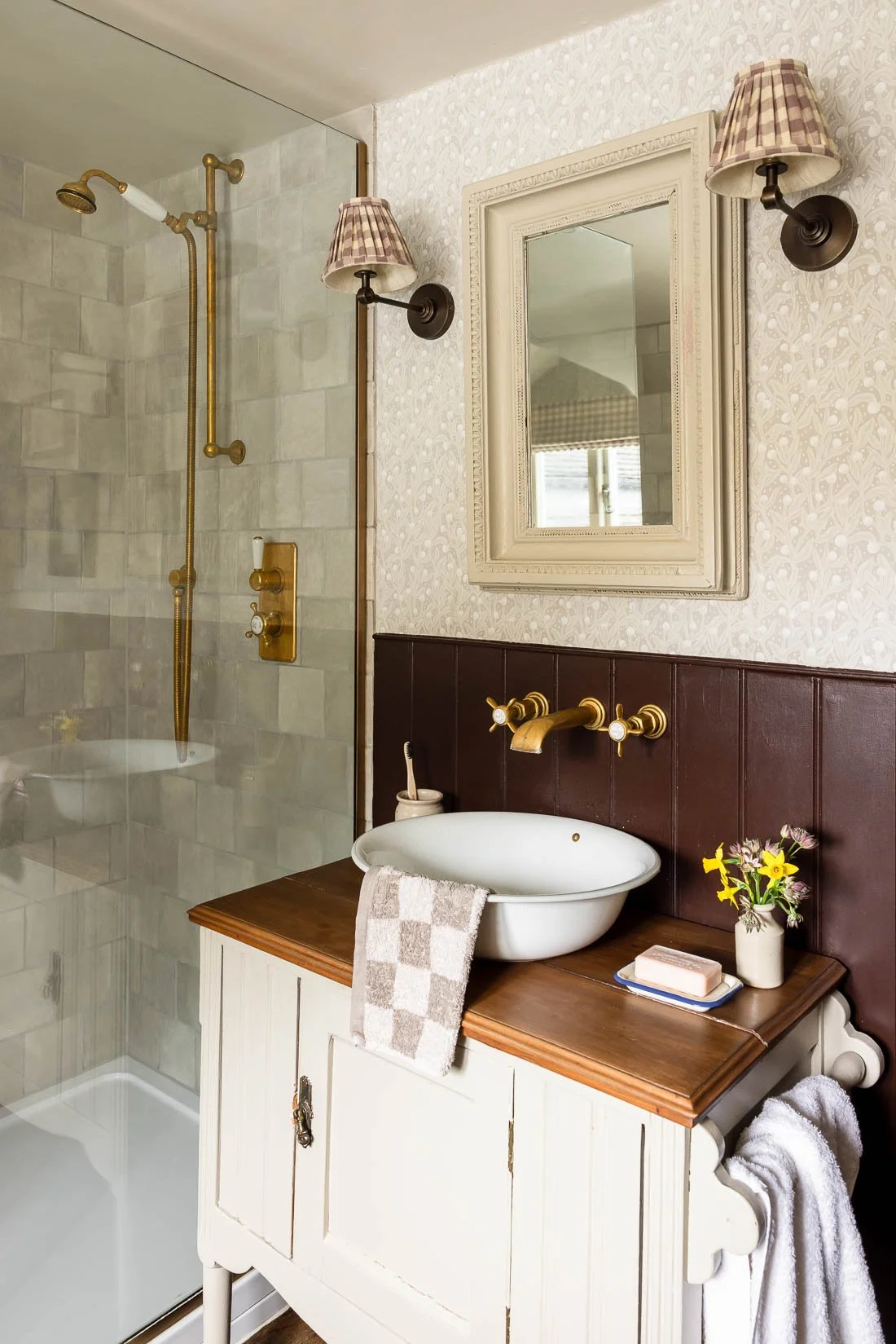 A bathroom with a shower stall, a vintage style basin sink, a mirror, floral wallpaper, wall-mounted lamps, and a wooden cabinet with decorative items including a vase of flowers and soap.