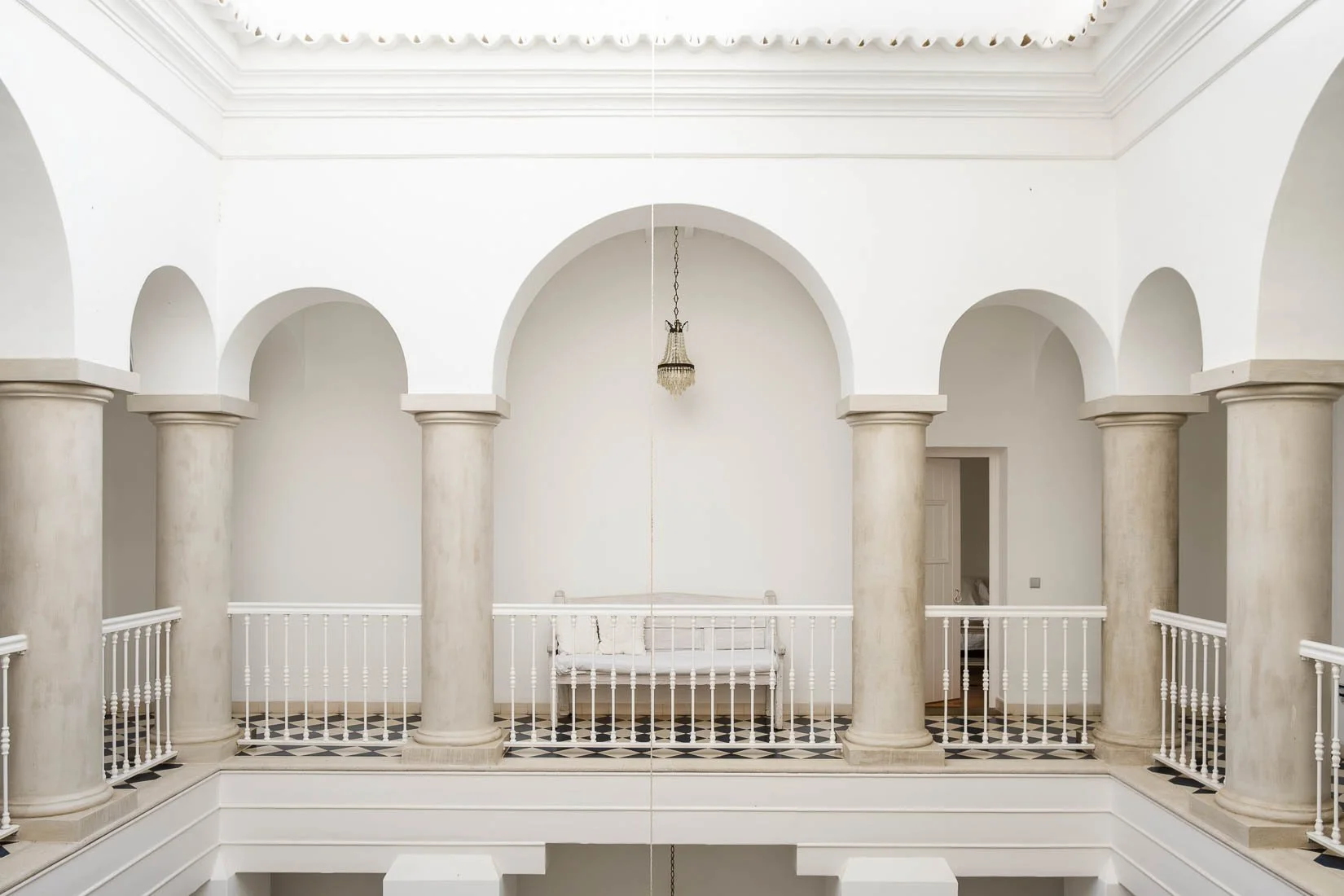 Enclosed balcony with four large white columns, white railing, a small white bench, chandelier, and open doorway in a bright, white-walled interior.