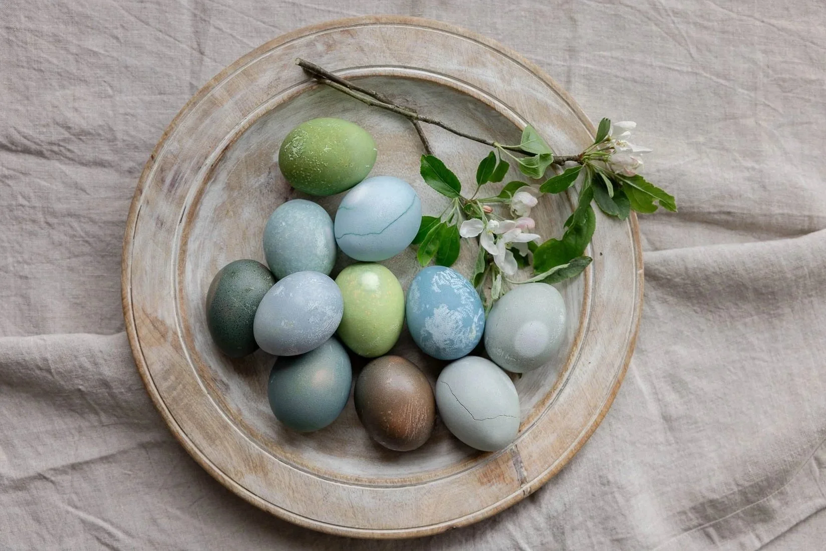 A round wooden plate containing pastel-colored eggs and a sprig of white flowers and green leaves placed on a beige fabric background.
