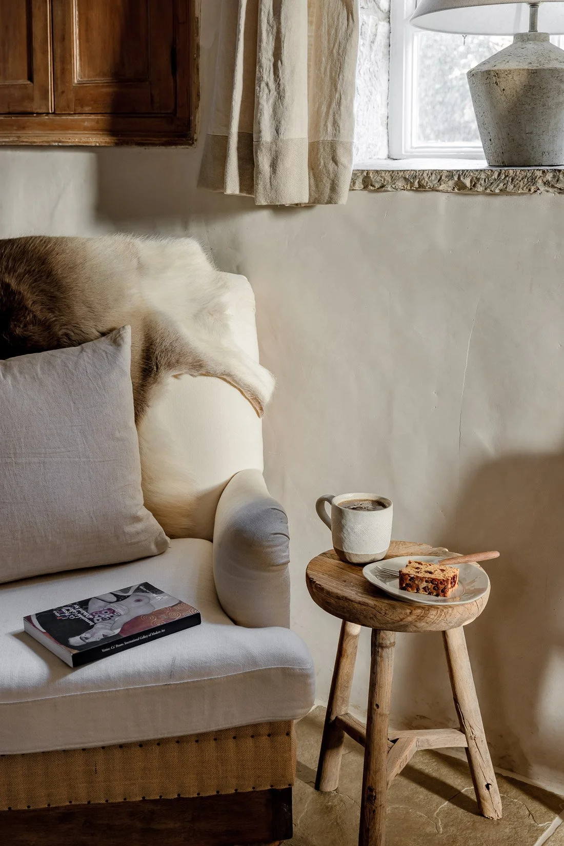 Cozy living room corner with a beige couch, a book, a mug, and a slice of cake on a wooden stool near a window with beige curtains.
