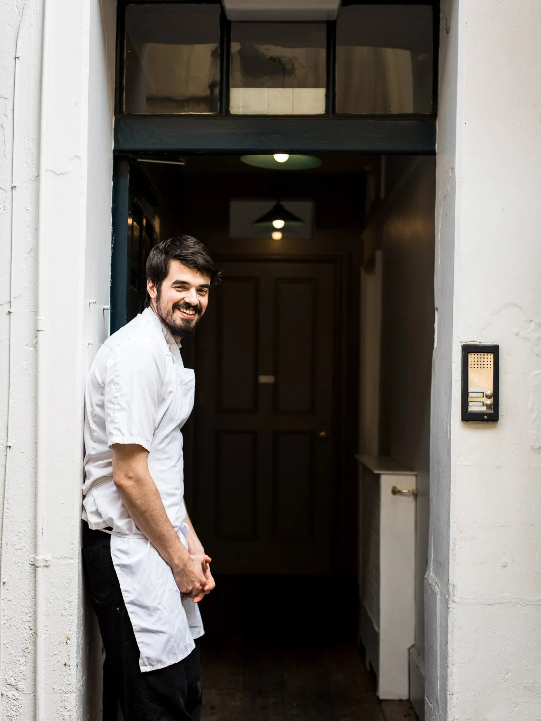 A smiling man in a white chef's coat standing in an apartment doorway.