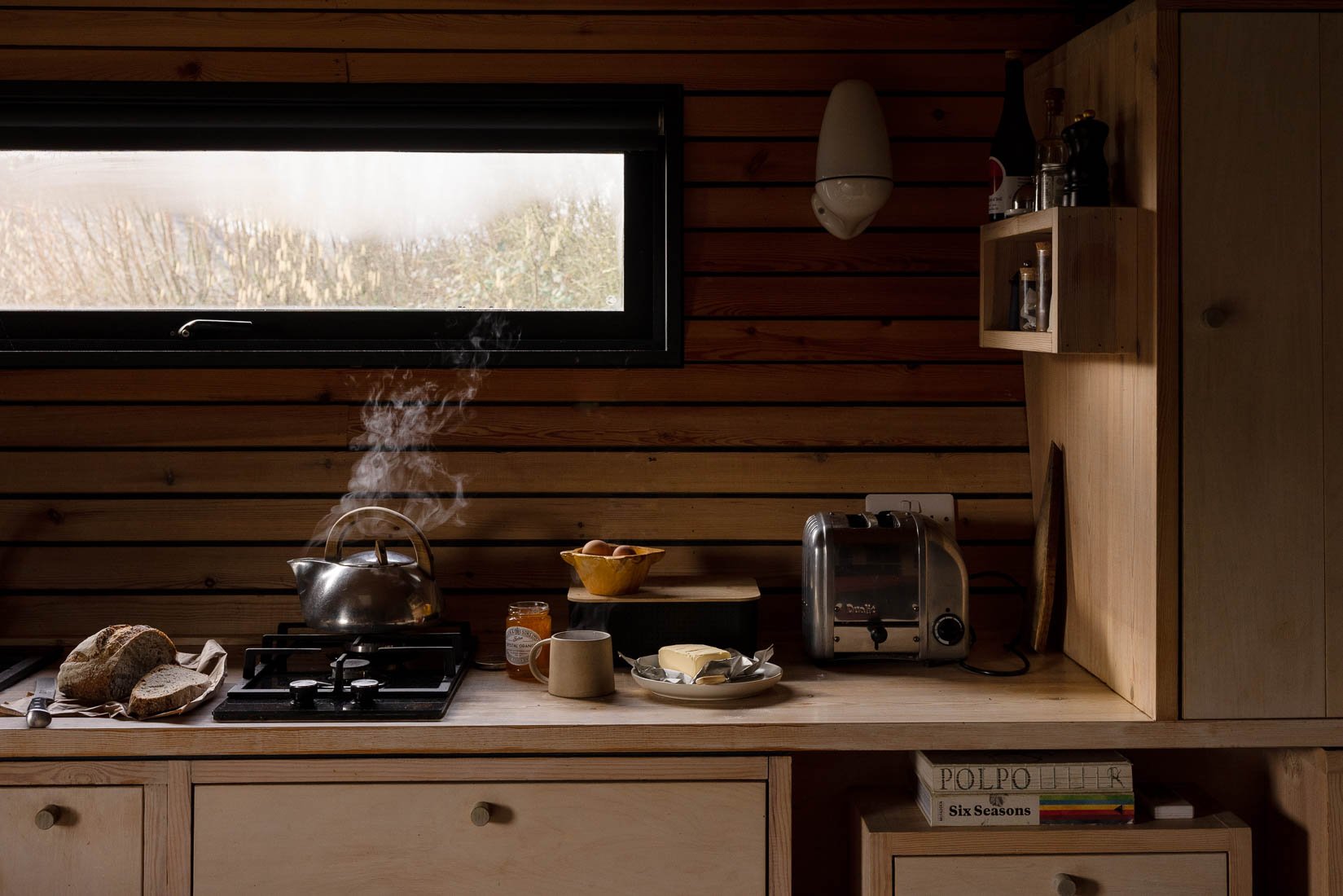 A rustic kitchen with wooden walls, a window, a stove with a steaming kettle, bread on a cutting board, a butter dish, a mug, a jar of jam, a bowl of eggs, a toaster, and a small shelf with bottles and jars.