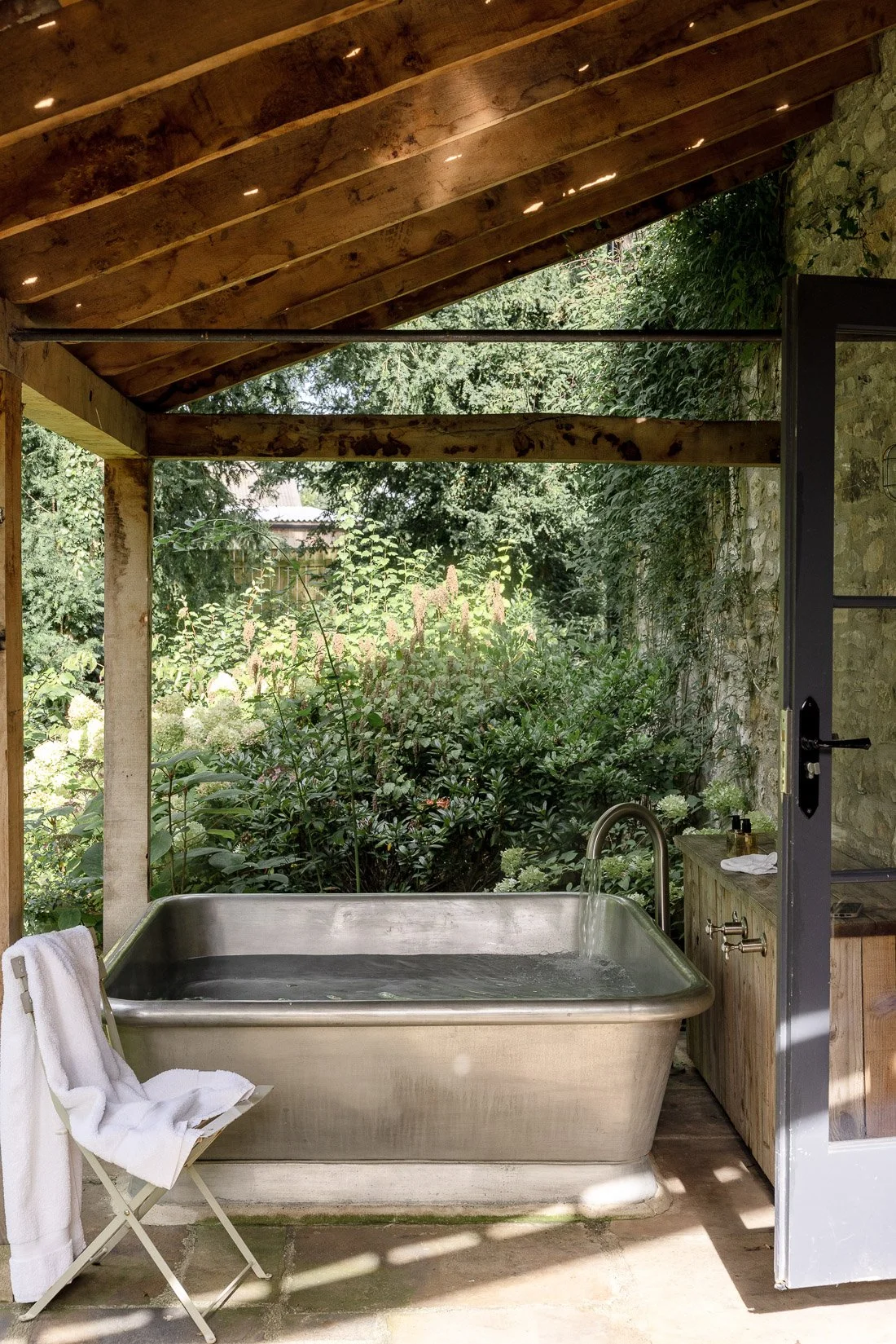 Outdoor rustic bathtub with water, a faucet, and a towel on a chair, surrounded by greenery and a stone wall, under a wooden roof.