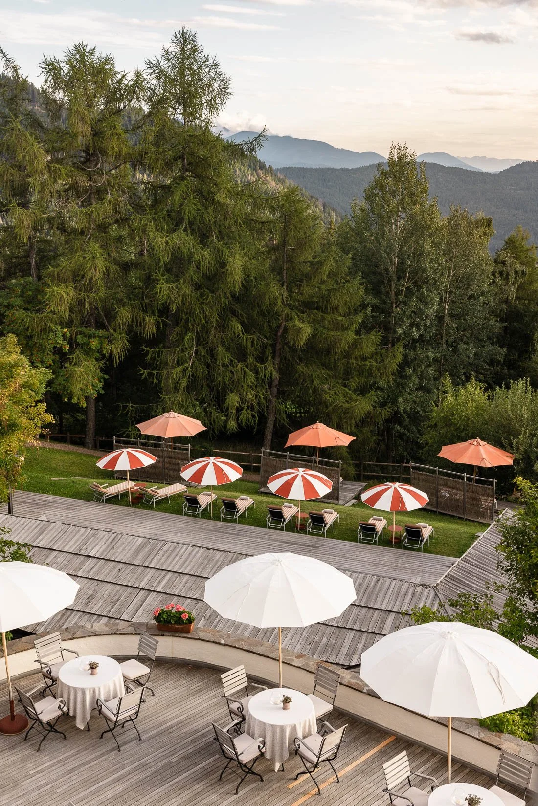 Outdoor patio area with round tables, white tablecloths, and white umbrellas, overlooking a grassy yard with red and white striped umbrellas, lounge chairs, tall trees, and mountain scenery in the background.