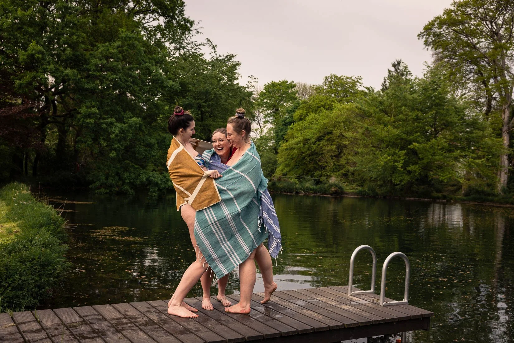 Three women smiling and hugging on a dock by a lake, wrapped in blankets with trees in the background.