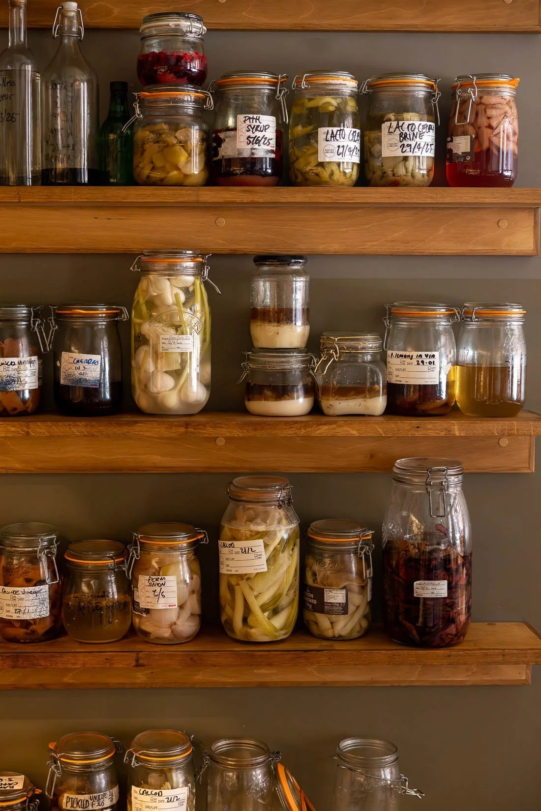 Shelves with glass jars containing pickled vegetables, fruits, and liquids, labeled with dates and contents, in a pantry or kitchen.