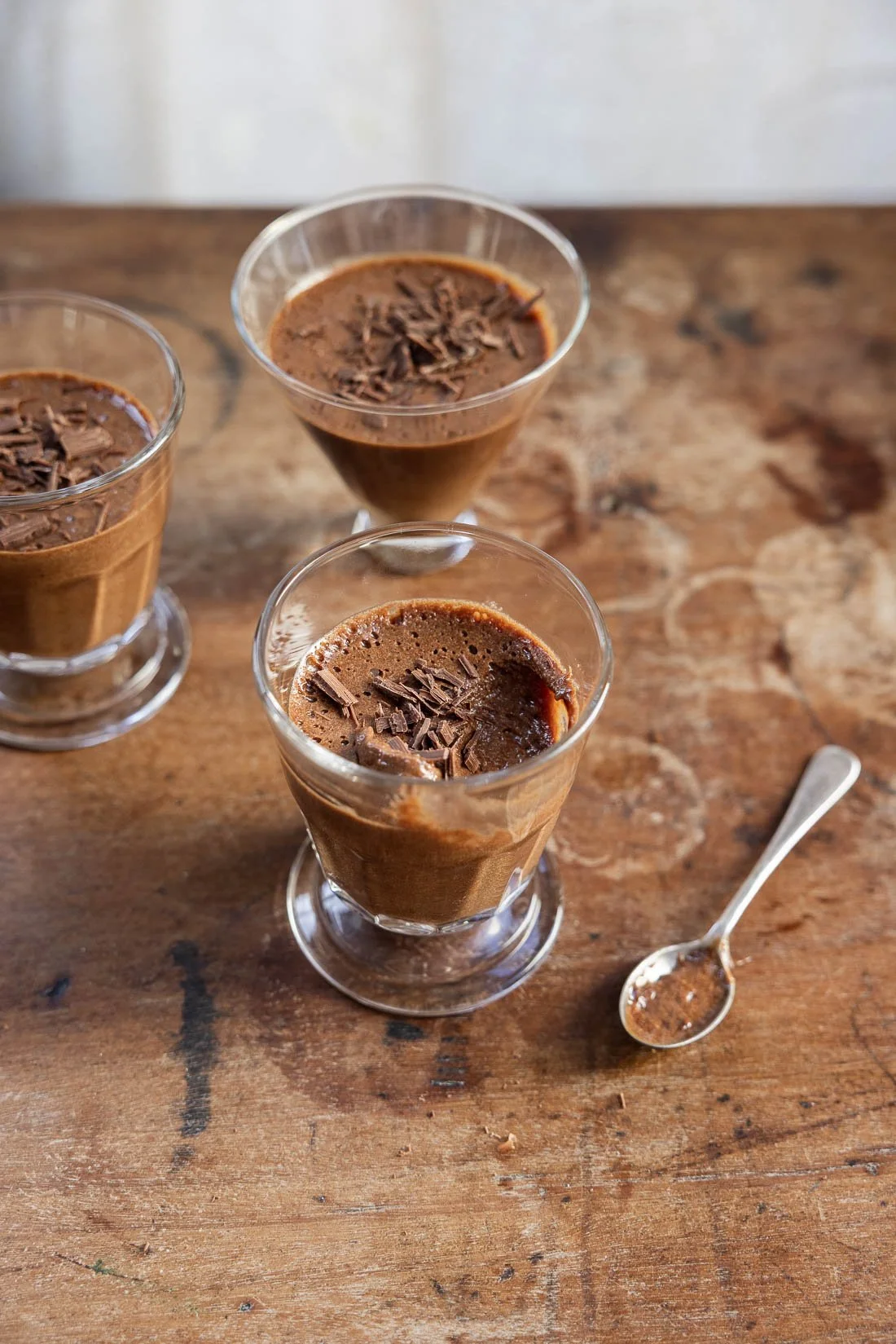 Three glass cups of chocolate mousse topped with chocolate shavings on a wooden table, with a silver spoon beside one of the glasses.