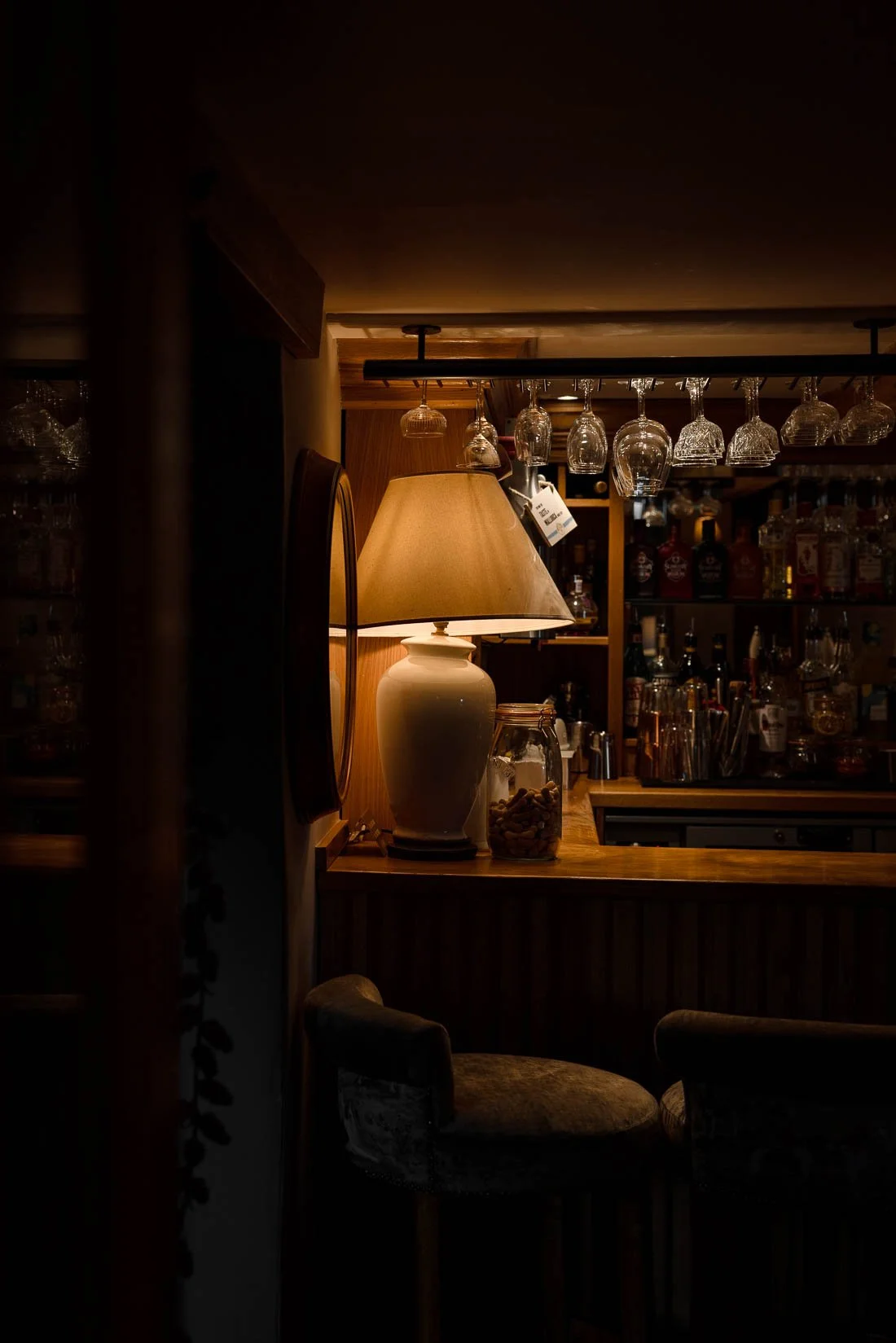 A cozy bar scene featuring a warm table lamp, hanging glassware, and shelves stocked with bottles, set against a dark background.