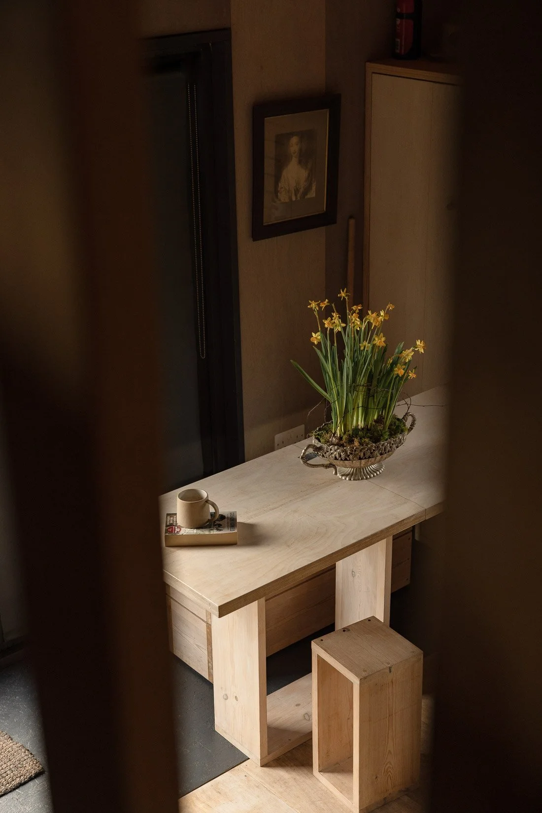 A wooden table with a potted plant with yellow flowers, a coffee mug, and a magazine, set against a dark wall with a framed portrait of a woman.