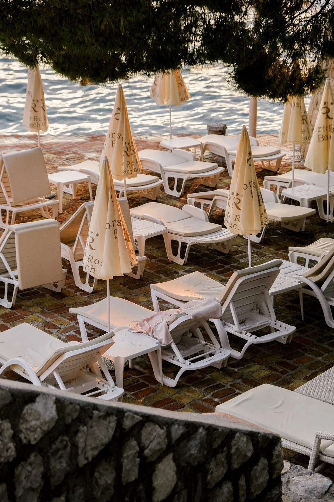 Empty white lounge chairs and umbrellas on a brick patio by the water, with trees overhead and sand in the background.