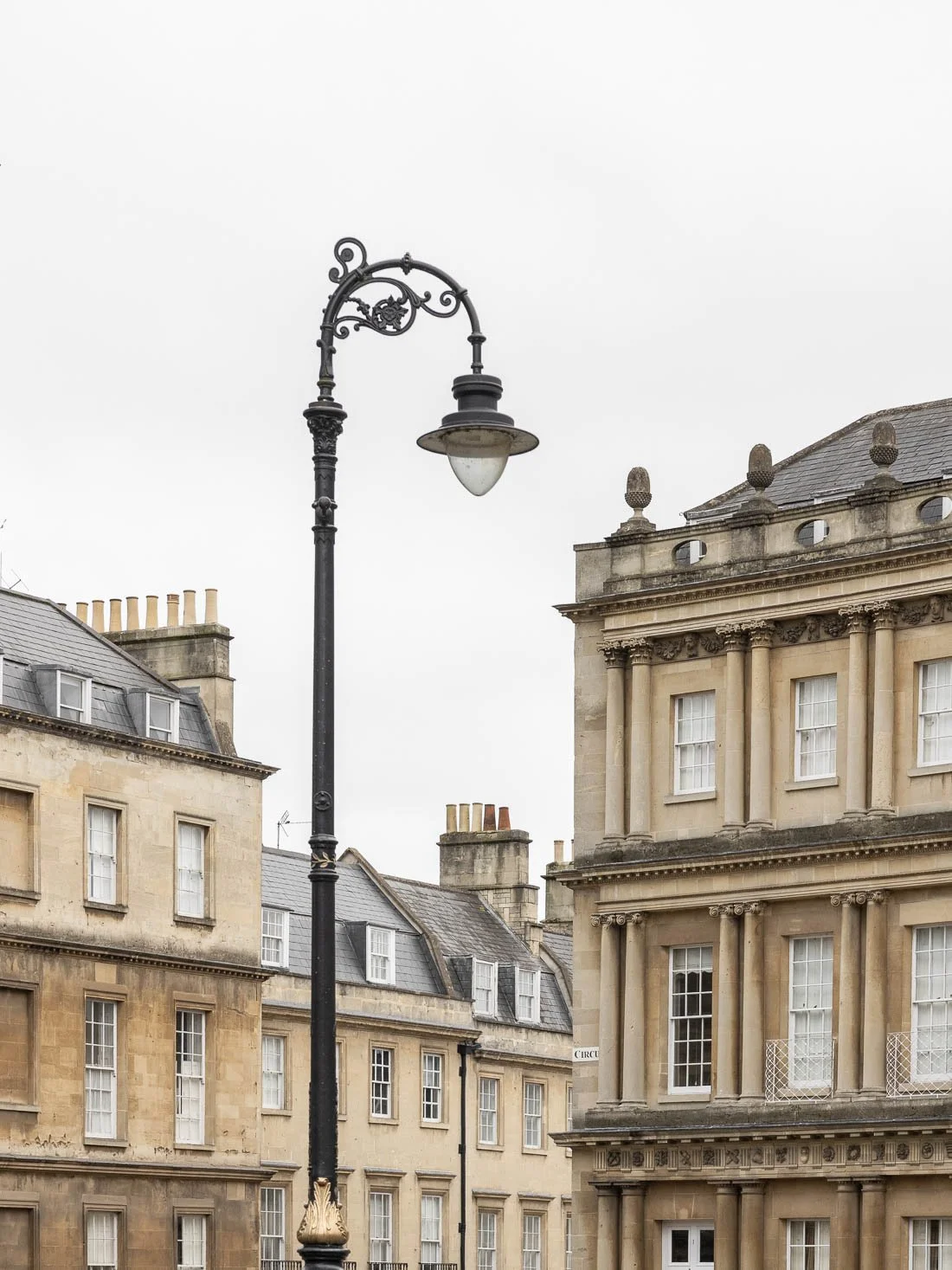 A vintage black street lamp in front of historic stone buildings with multiple windows and decorative architectural details, under a cloudy sky.