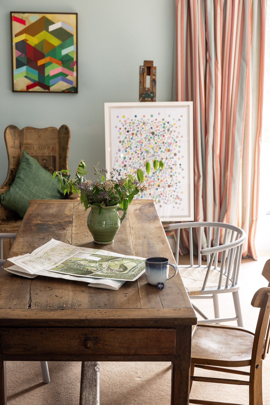 A rustic wooden dining table with documents and a ceramic mug, a green vase with flowers, a gray chair, a wooden chair, a sofa with a green pillow, colorful abstract artwork on the wall, a framed dot pattern art piece, and striped curtains in a brigh