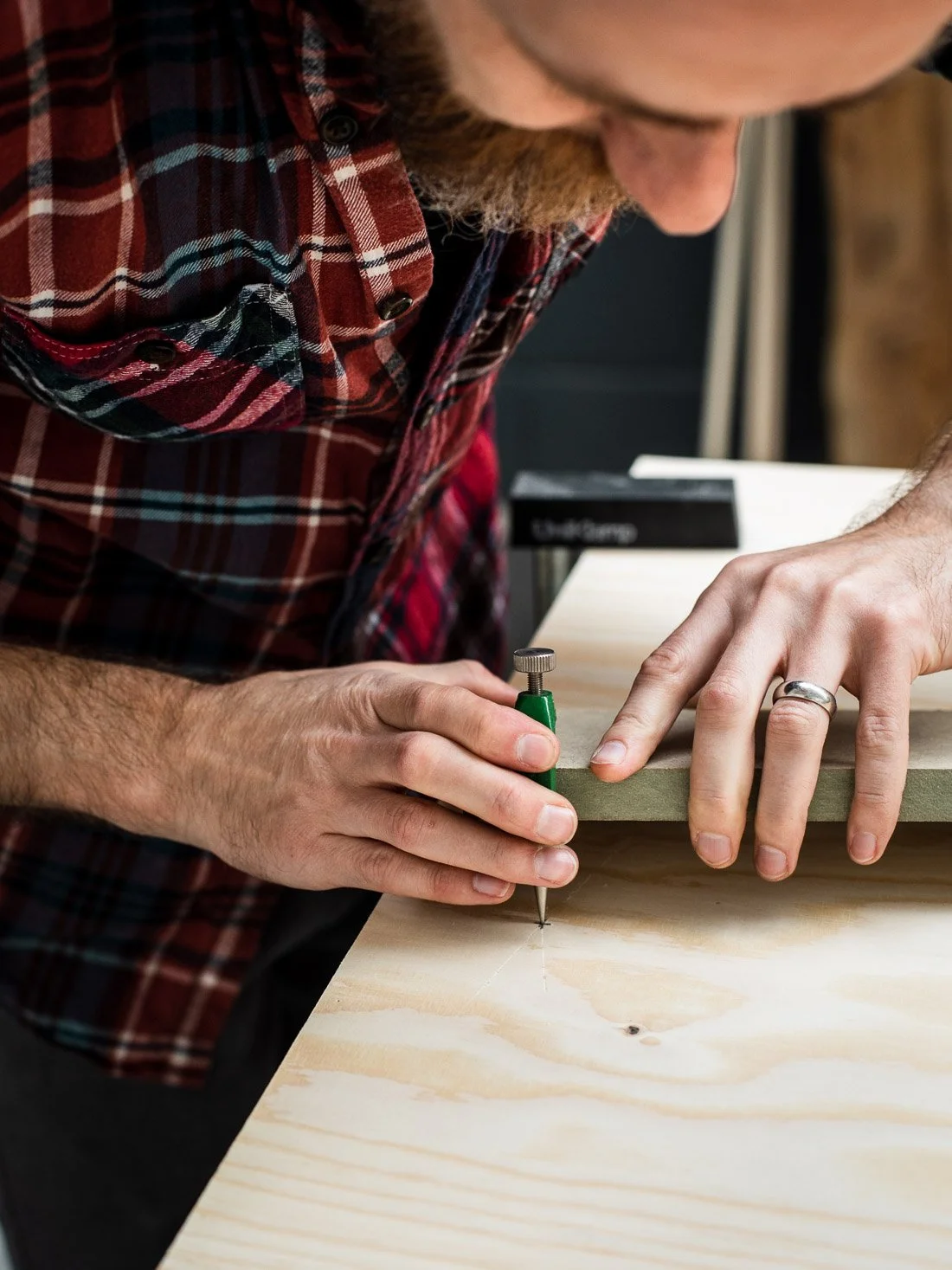 A person wearing a red and black plaid shirt using a green marking gauge to mark a wooden board.