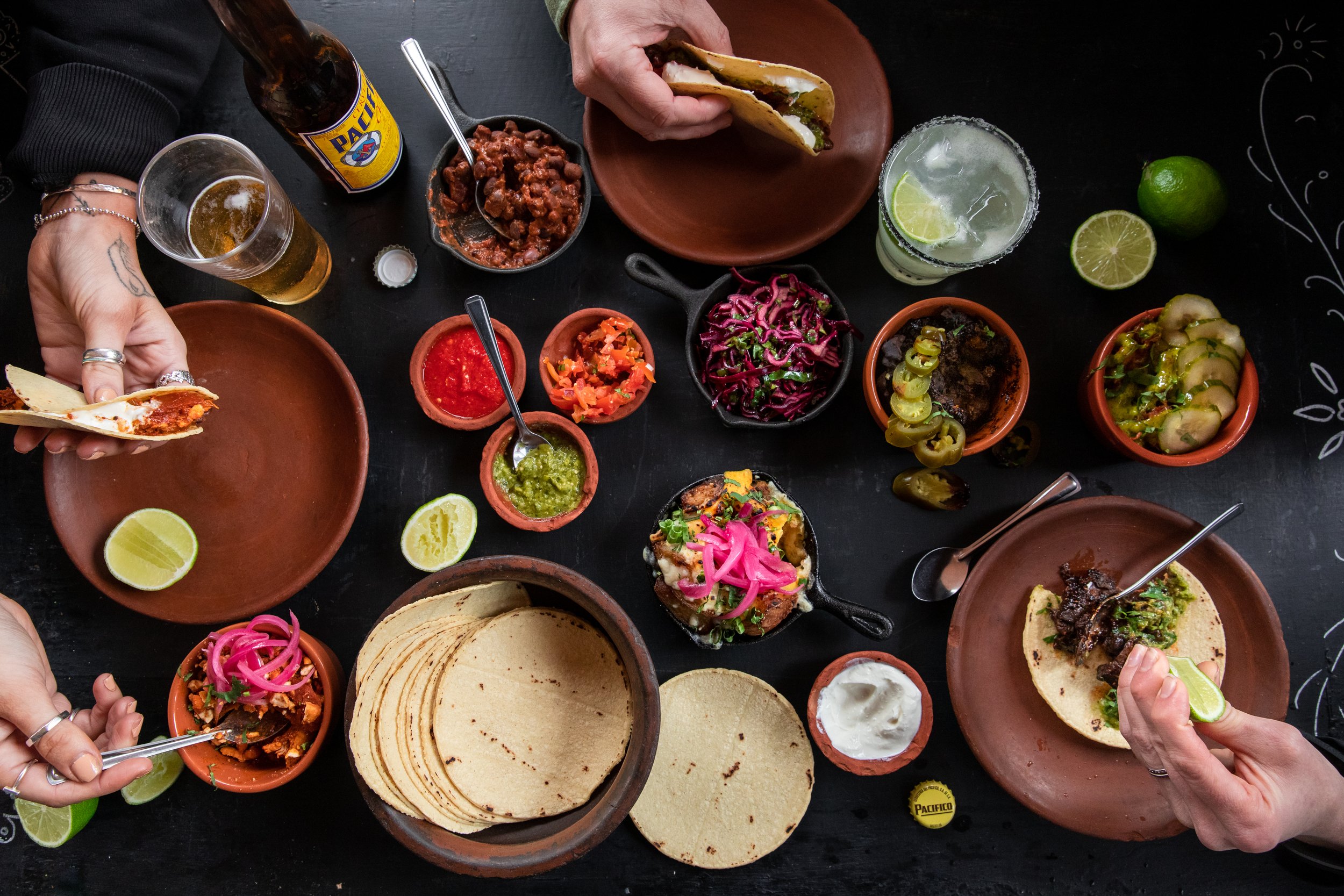 People enjoying Mexican food with tacos, salsa, chips, and beverages on a black table, with lime wedges and various bowls of toppings.