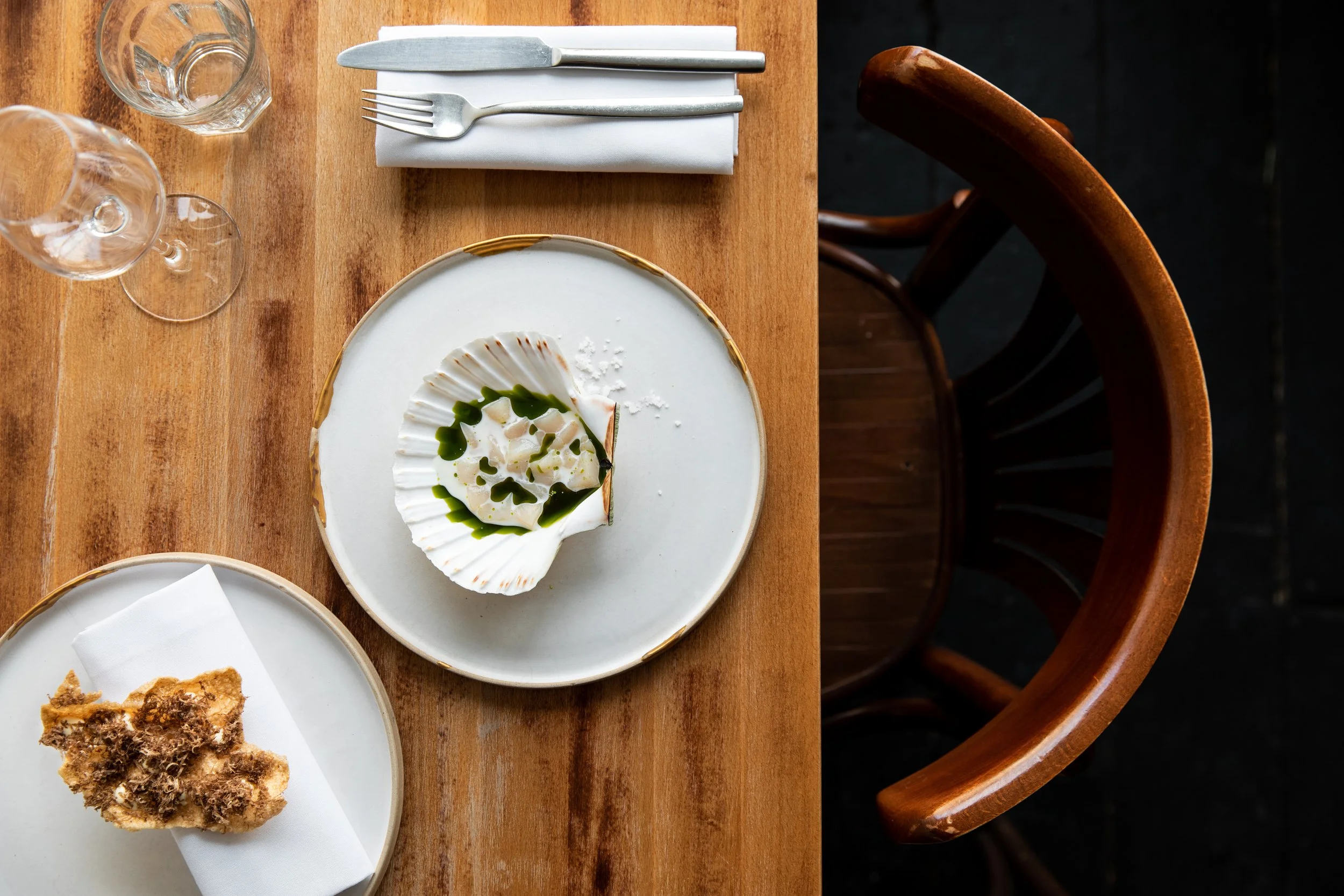 A wooden dining table set with a plate of scallops topped with green sauce, two glasses of water, a folded white napkin with a knife and fork, and a plate of fried food. A dark wooden chair is positioned next to the table.