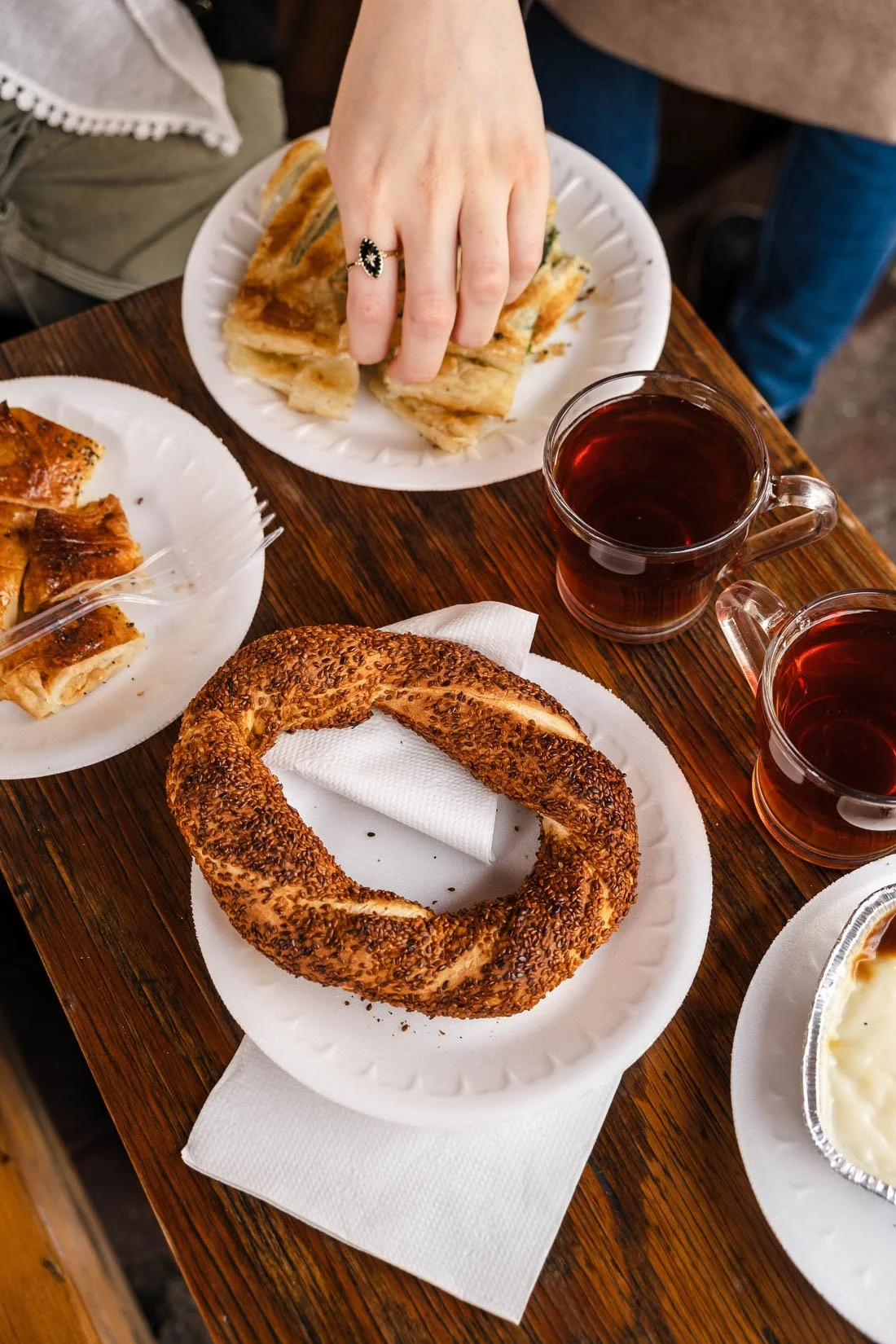 A table with Turkish-style foods, including a sesame-covered bagel, sliced flatbreads on paper plates, and two glasses of tea.