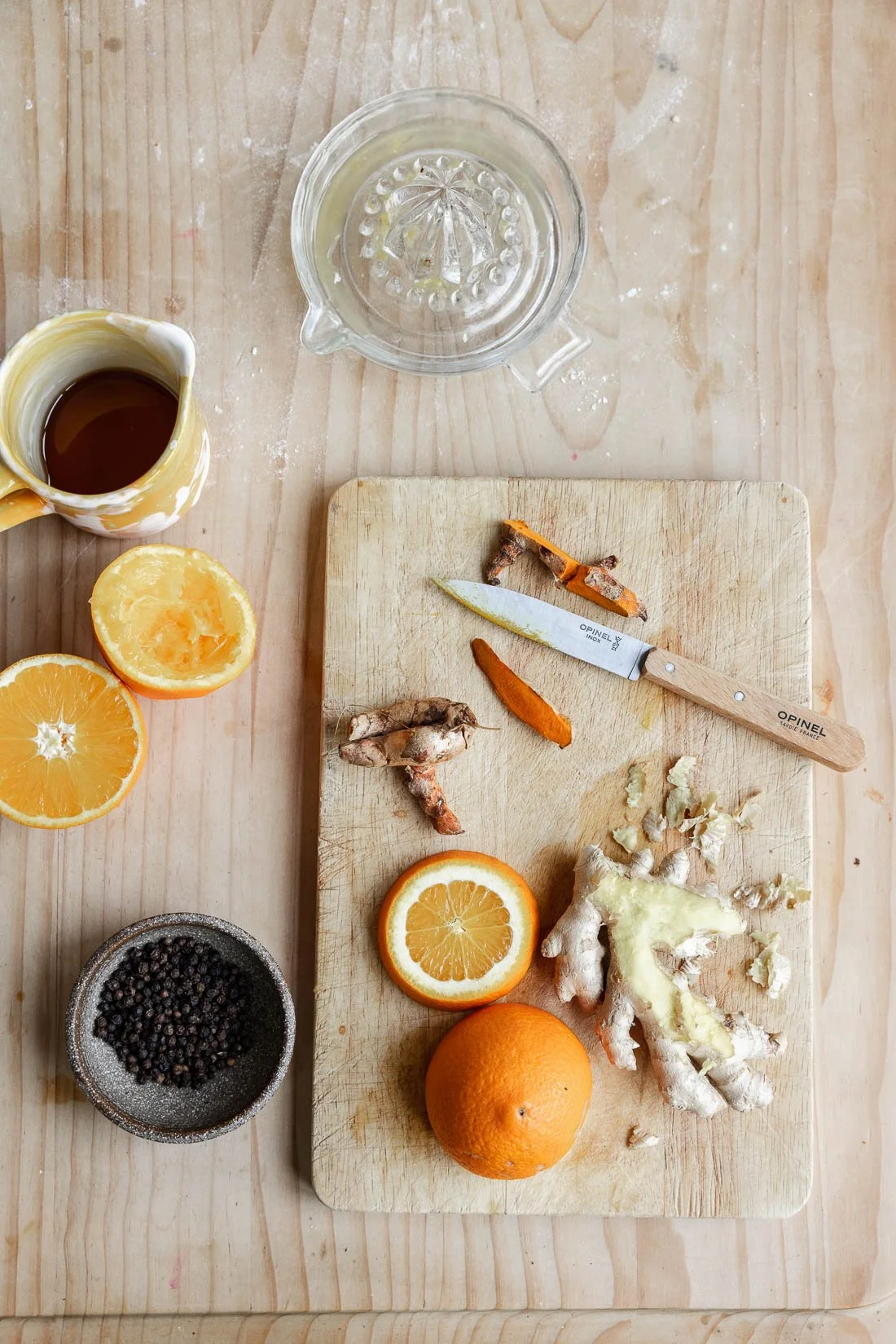A wooden cutting board with sliced oranges, turmeric, ginger, and ginger root, along with a knife, surrounded by a glass lemon juicer, a small pitcher of juice, a bowl of peppercorns, and half a lemon on a wooden table.