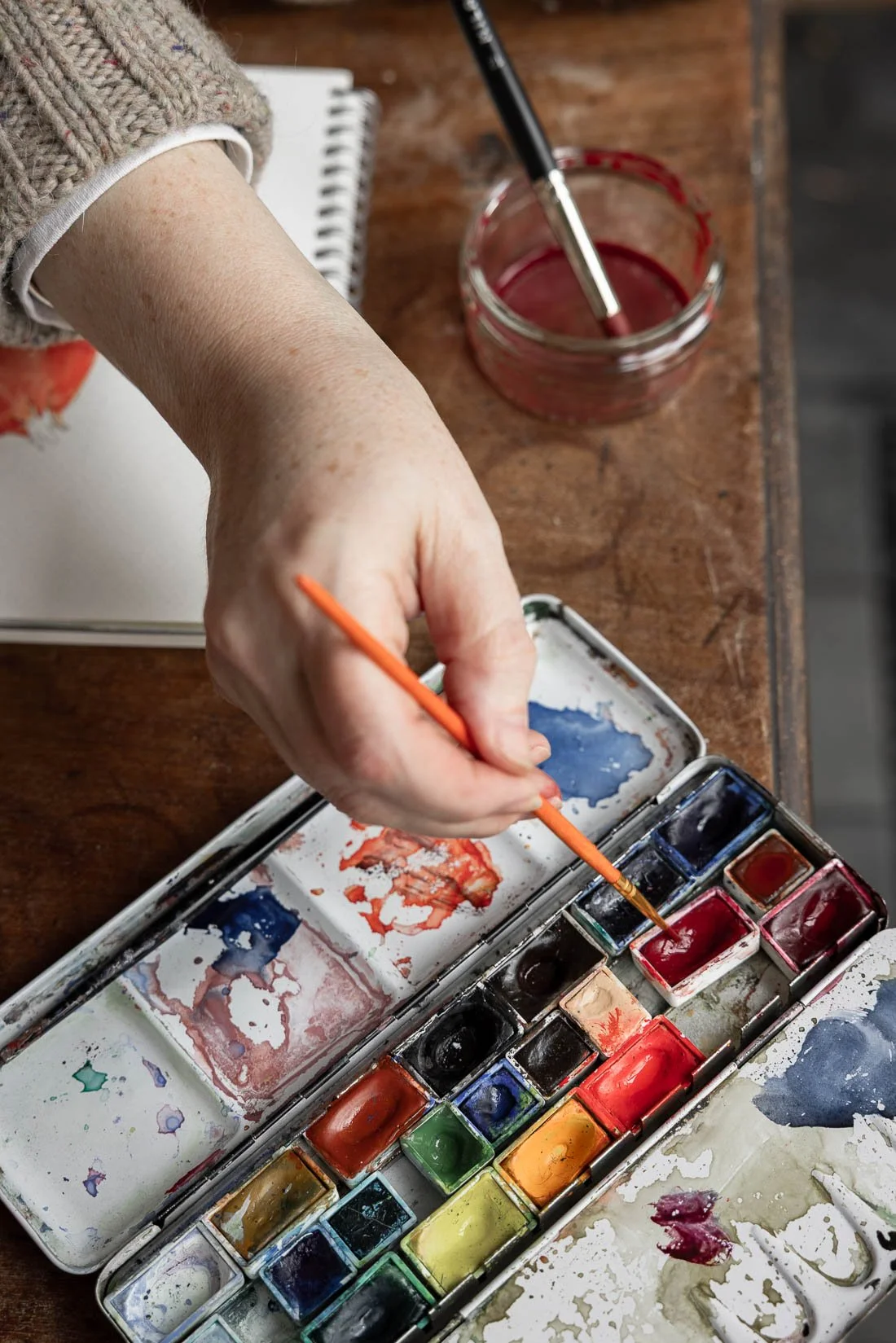 A person painting with watercolors, using an orange paintbrush dipped in red paint. The watercolor palette includes multiple colors, some mixed or used. The person is wearing a textured, knitted sweater, and the workspace is a wooden table, with a sm