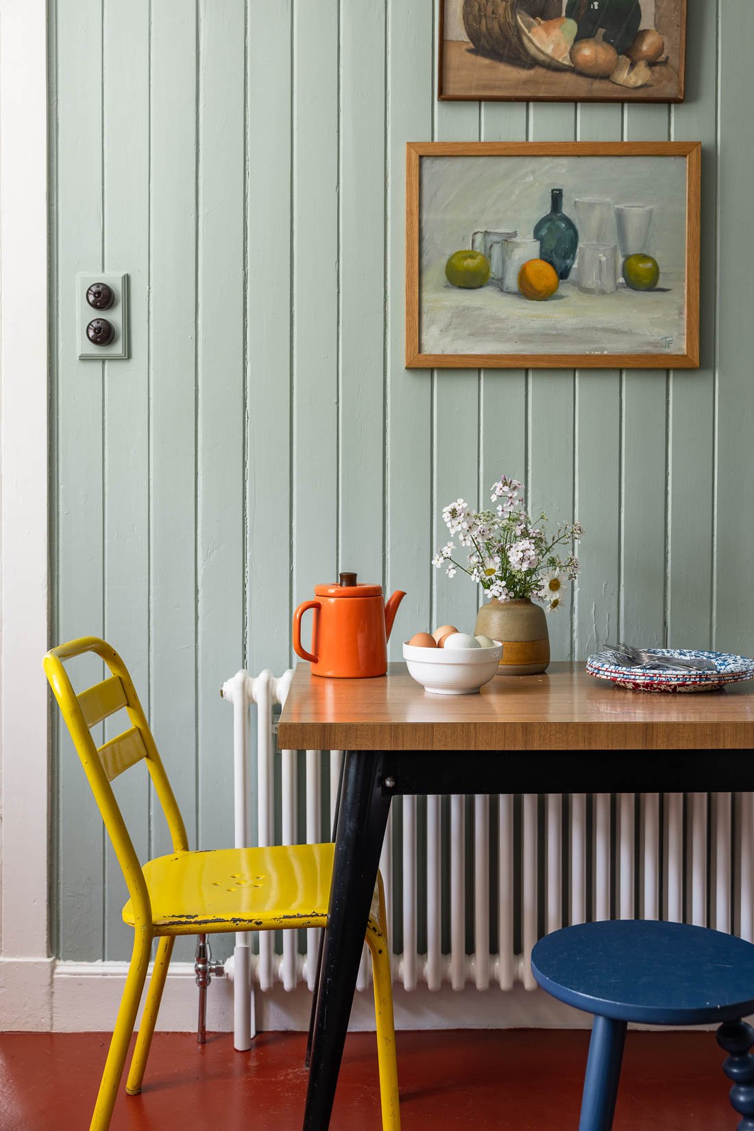A dining area with a wooden table and three chairs, one yellow, one black, and one blue. On the table are a white bowl of eggs, an orange teapot, a small beige pot with white flowers, and a colorful plate. The wall behind is light green with vertical
