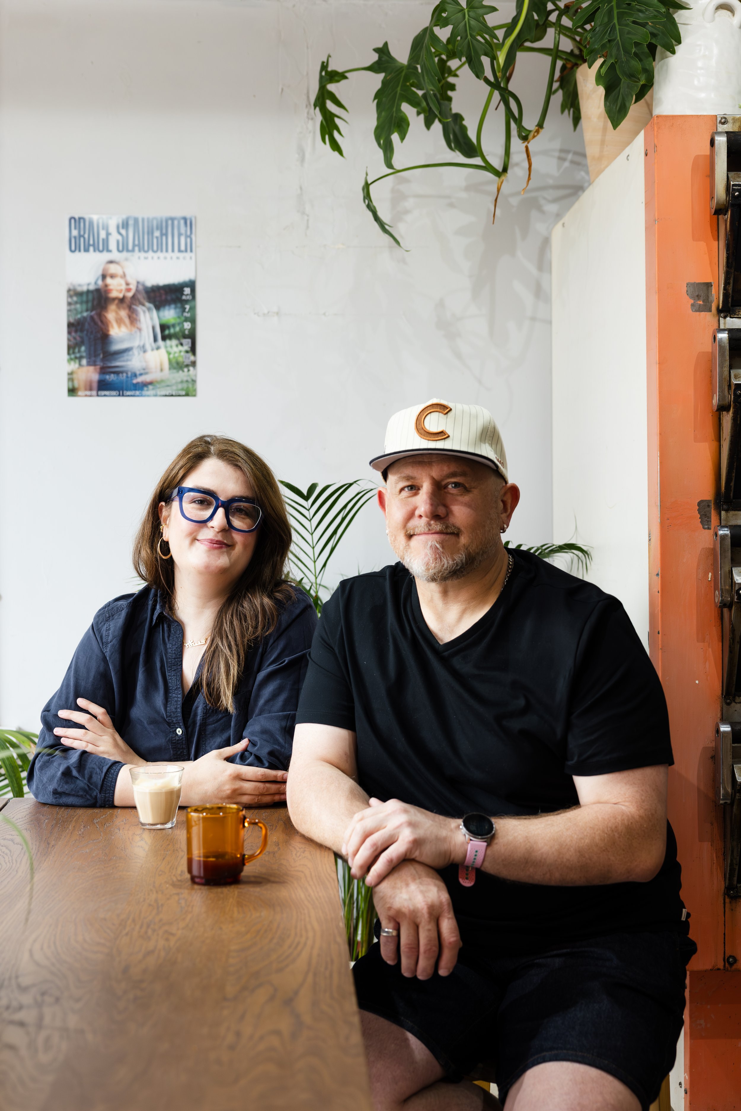 Two people sitting at a wooden table, a woman with long brown hair wearing glasses and a dark shirt, and a man with a gray beard wearing a white cap with a brown 'C' and a black shirt. There are two drinks on the table, one in a clear glass and the o