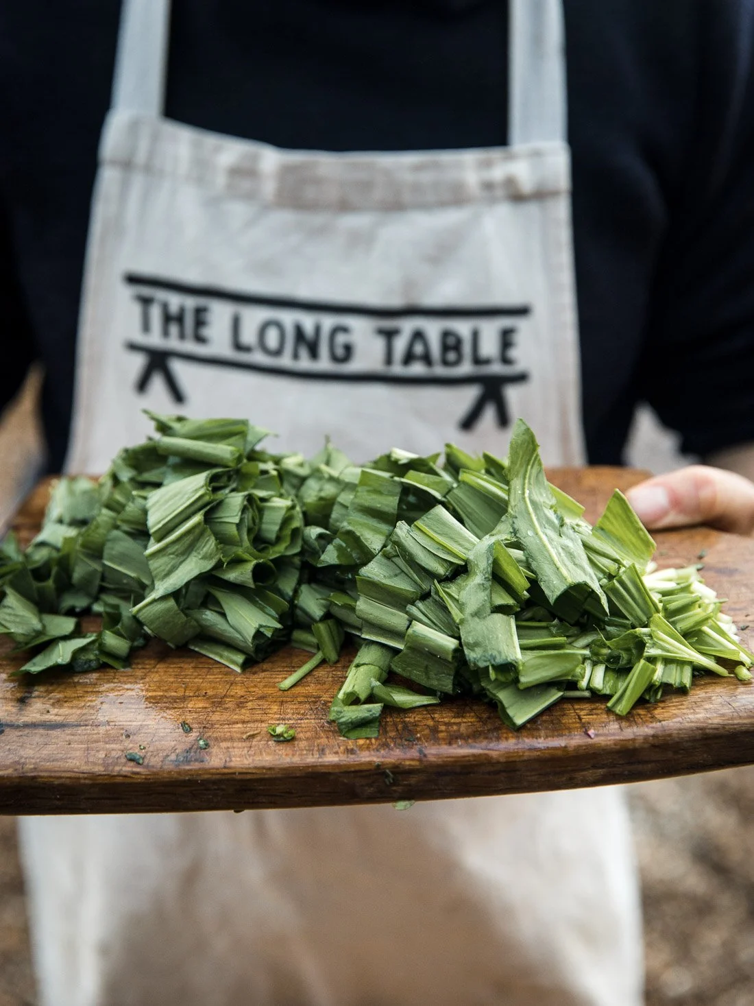 Person holding a wooden board with chopped green vegetables, wearing a white apron with 'The Long Table' written on it.