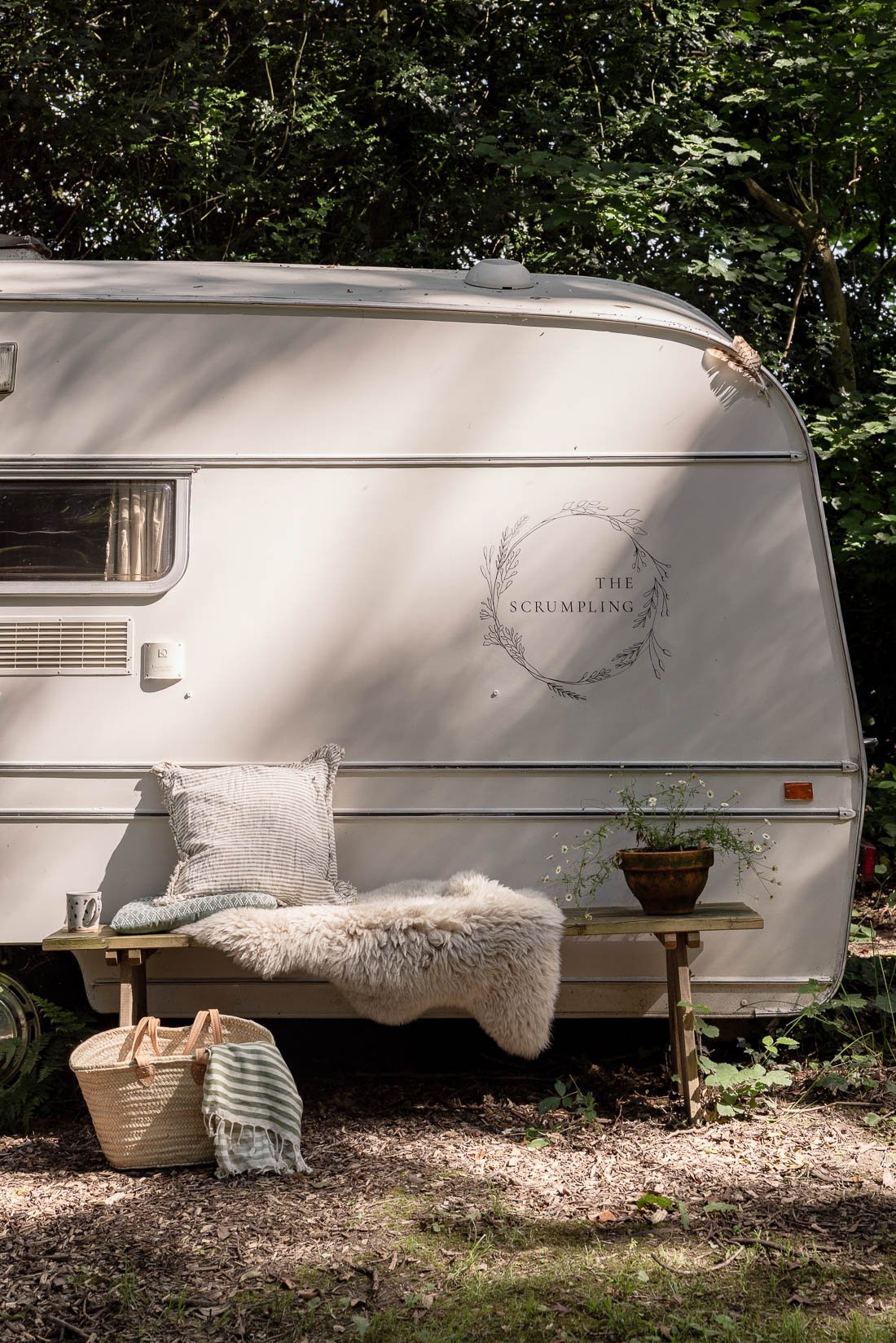 A white vintage camper trailer with a decorative wreath graphic and the words "The Scrumbling" on the side, set in a wooded outdoor area. A small wooden bench in front of the camper holds a pillow, a sheepskin throw, and a potted plant. A woven baske