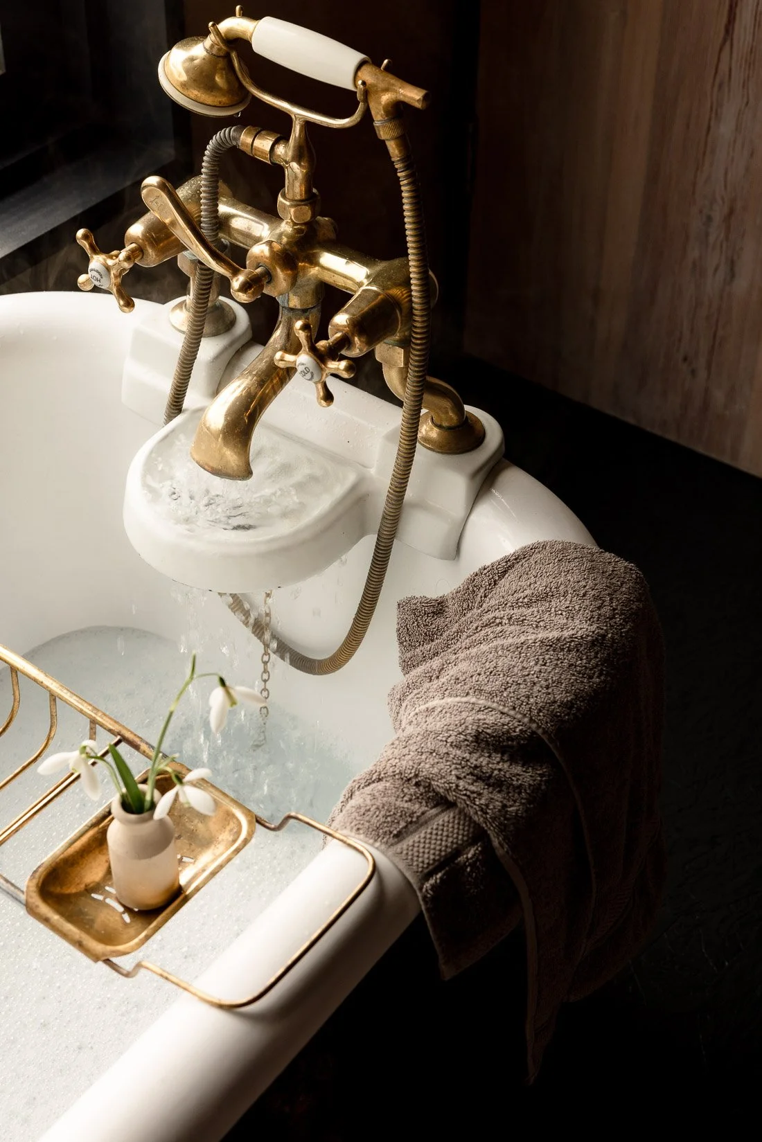 Vintage brass faucet with attached handheld shower head, water running into a white bathtub, with a brown towel and a small vase of white flowers nearby.