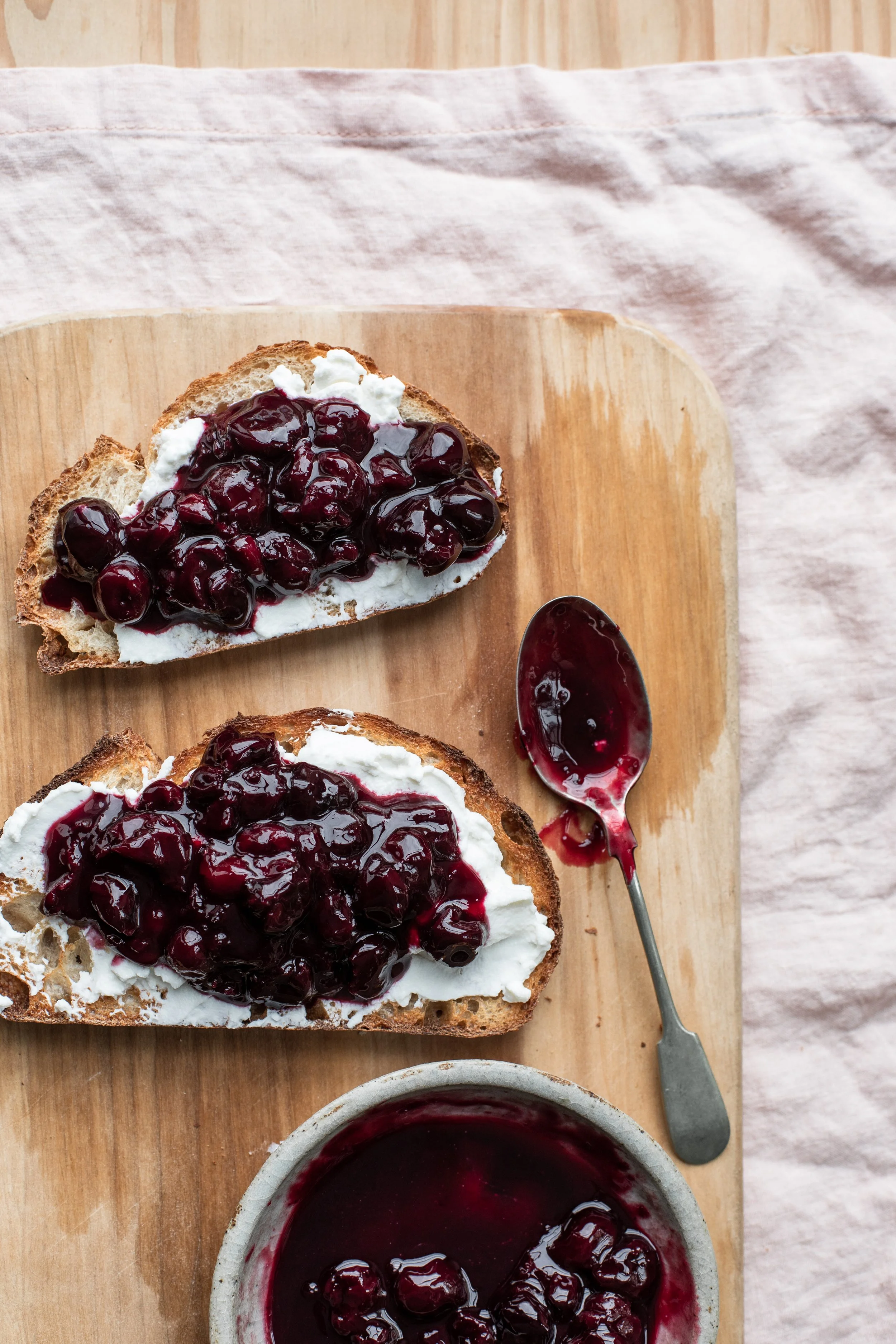 Sliced bread with cream cheese and cherry or blueberry jam, a spoon with jam, and a bowl of jam on a wooden surface.