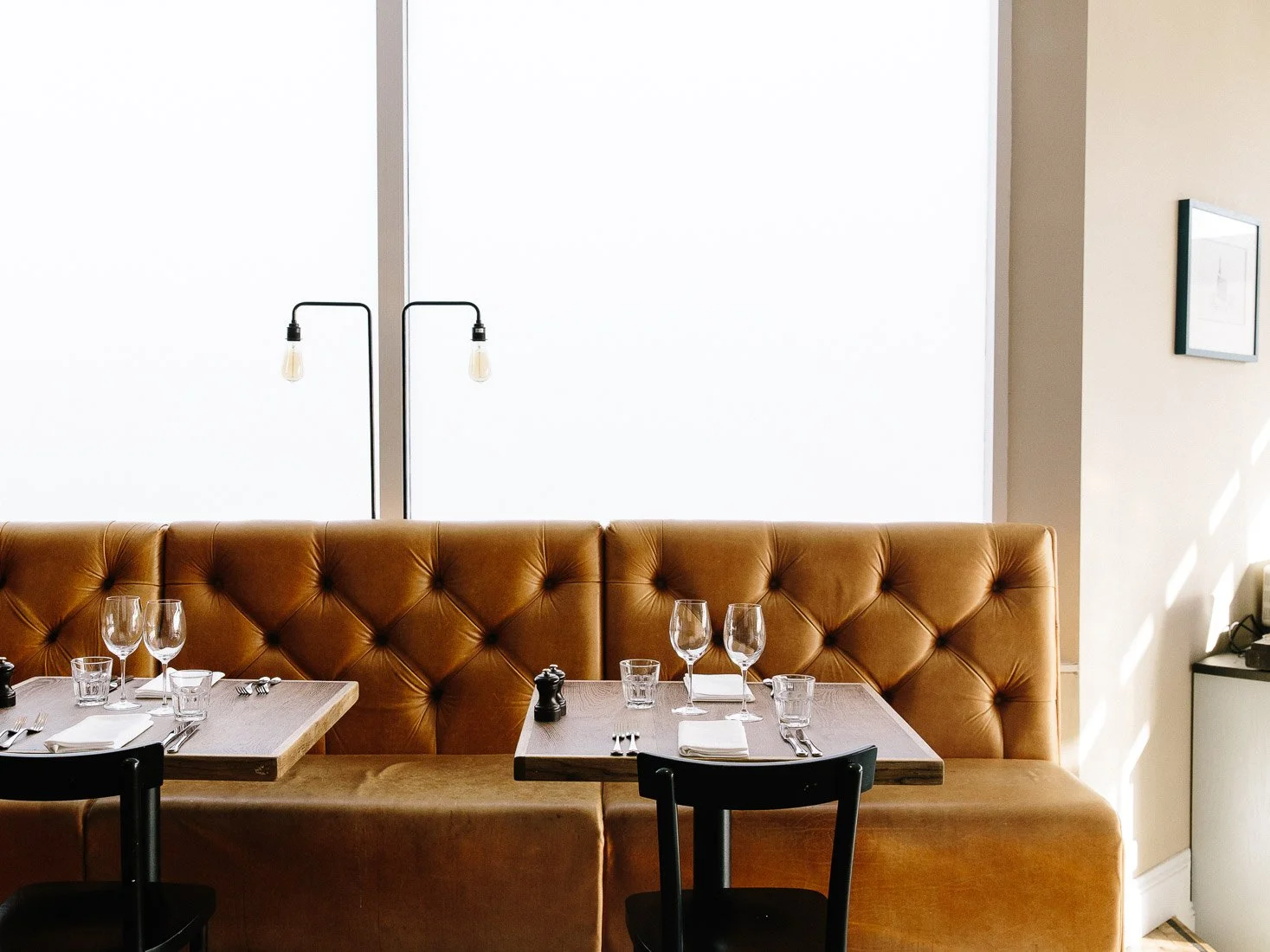 Restaurant booth with a leather cushioned back and a wooden table set with glassware, silverware, napkins, small candles, and a pepper mill, next to a large window with bright light.