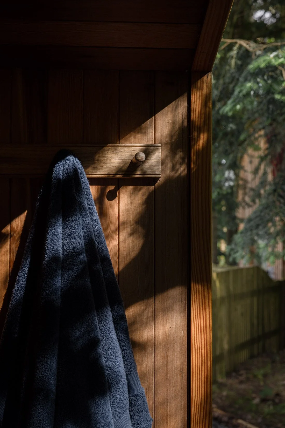 A wooden outdoor coat rack with a black fleece jacket hanging on it, illuminated by sunlight, with trees and a wooden fence in the background.