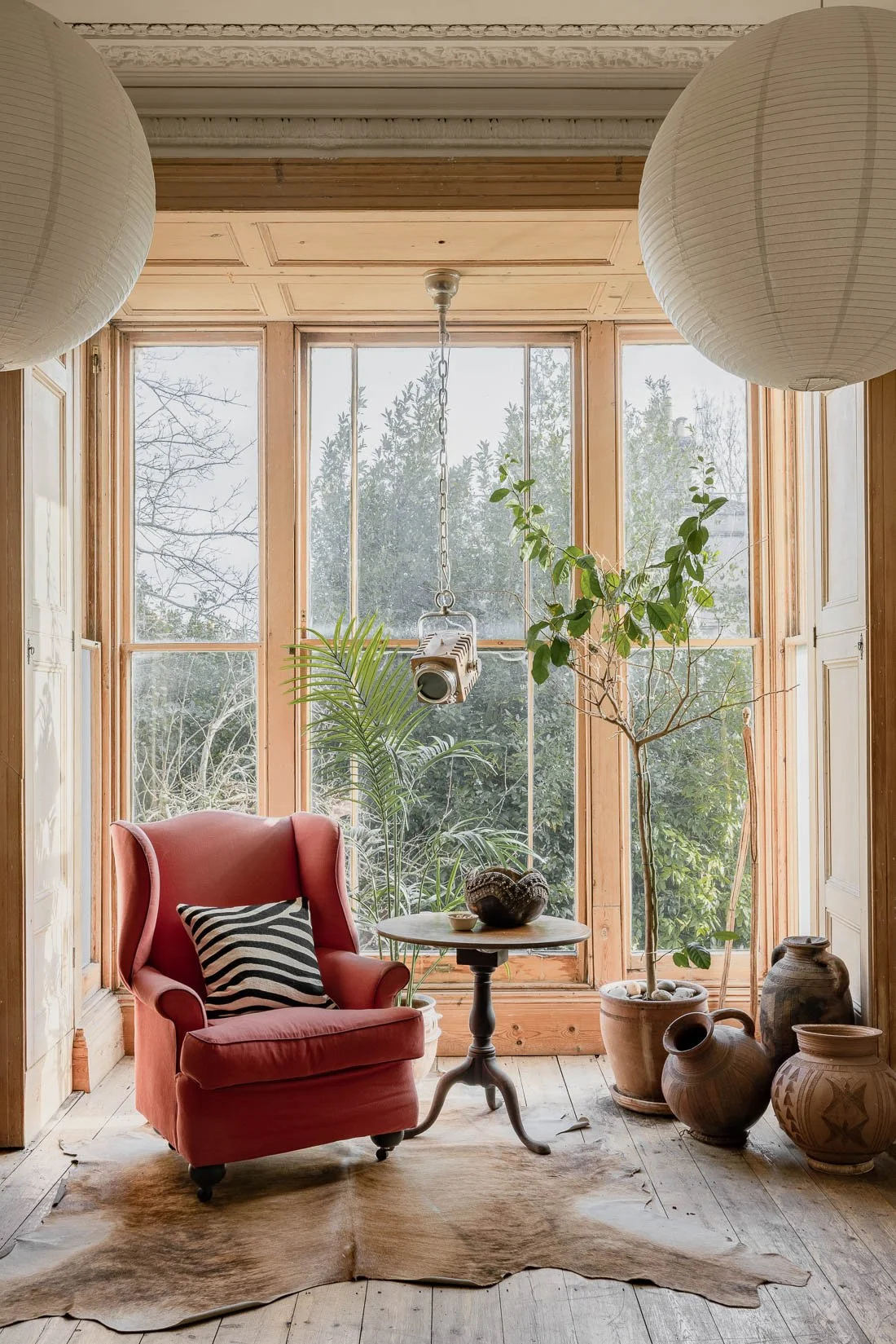 A cozy sunlit sitting area with a pink armchair, a striped pillow, a round table with decorative items, potted plants, and large windows revealing trees outside. There are traditional pottery vases on the floor and paper lanterns hanging from the cei