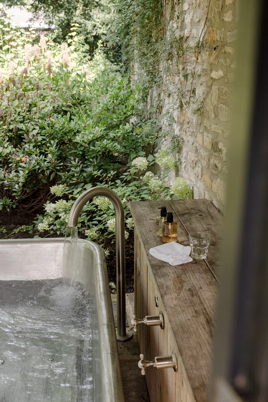 An outdoor bathtub in a garden with a wooden side table holding soap dispensers, a towel, and a glass of water. Green foliage and a stone wall are in the background.
