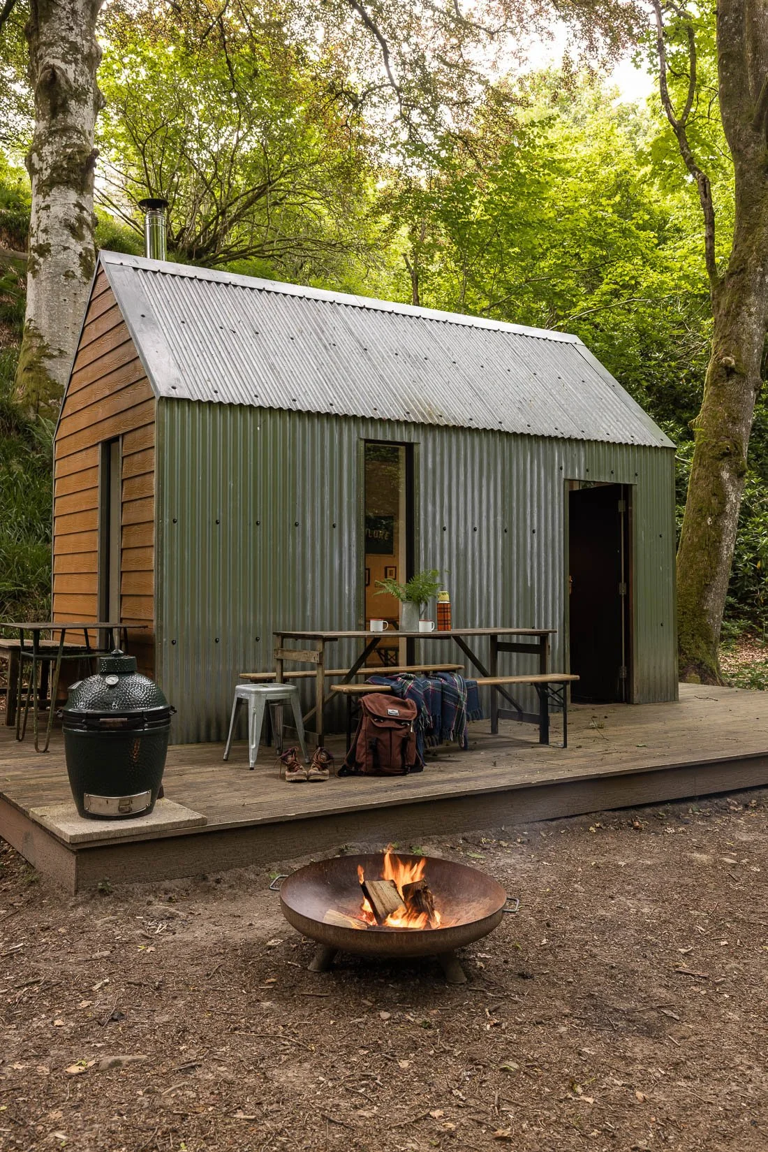 A small metal cabin with a corrugated metal roof, set in a forest. There is a wooden deck with outdoor furniture, a fire pit with burning wood, and trees surrounding the area.