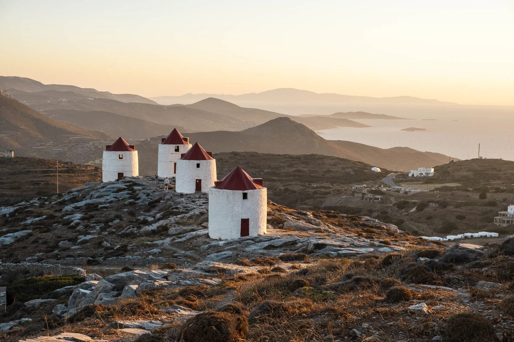 Four traditional white windmills with red conical roofs situated on rocky terrain overlooking a hilly landscape and a body of water in the distance, during sunset.