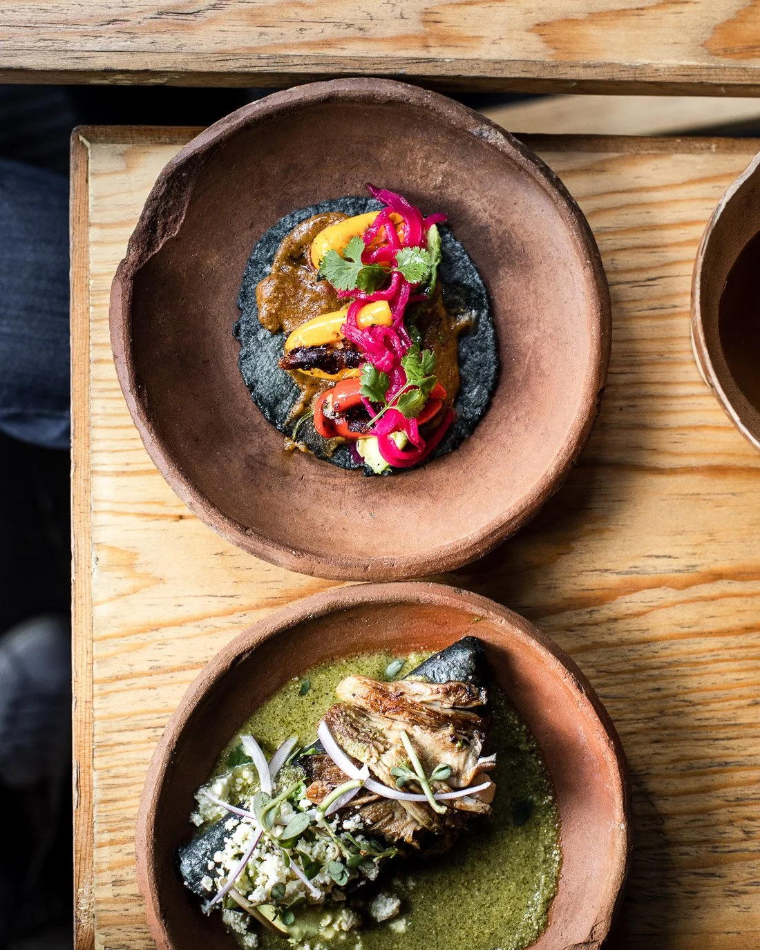 Two terracotta bowls with artwork on a wooden table; top bowl contains black tostada topped with colorful pickled vegetables and herbs, bottom bowl has a cooked fish fillet in green sauce with microgreens and cheese.