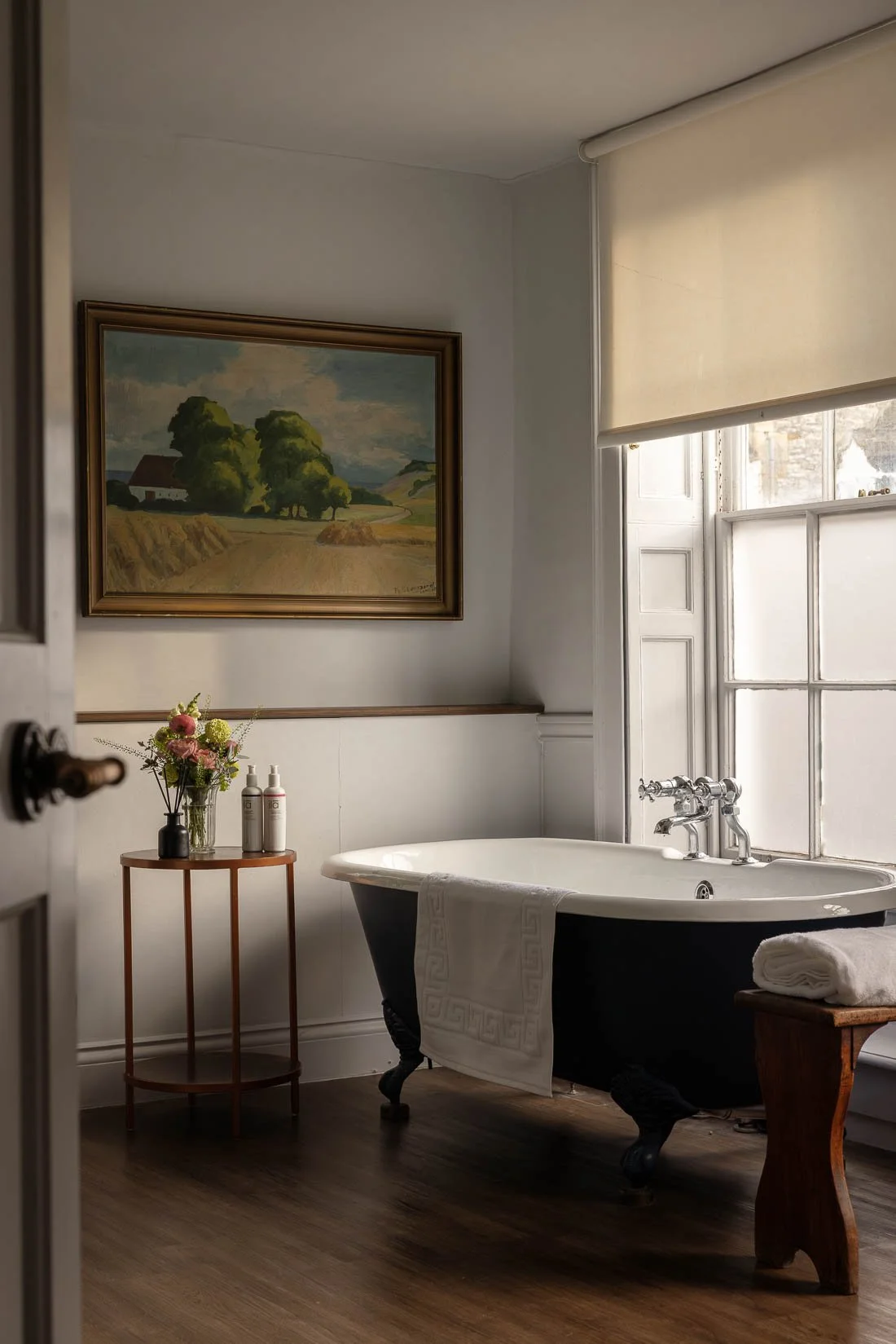 A vintage clawfoot bathtub with a white towel draped over the side, positioned near a window with frosted glass panes. Next to the tub is a small wooden table with rolled towels and a vase of flowers. On the wall above, there's a landscape painting o