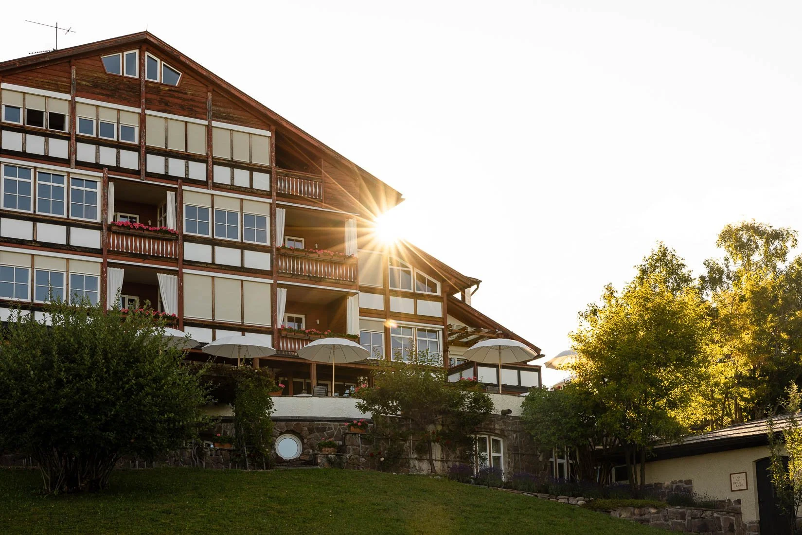 A multi-story building with wooden accents and balcony railings, under a bright setting sun with trees in the foreground.