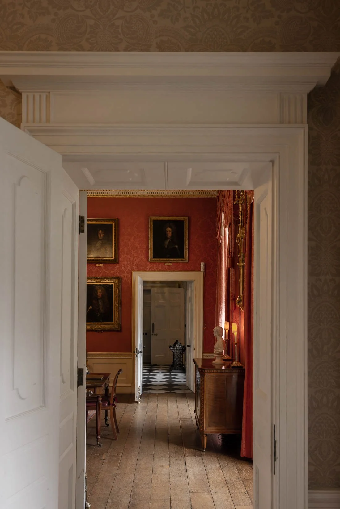 Interior view of a historic house with open doorways leading to a room with red walls decorated with framed portraits, a wooden side table with a bust sculpture, and a lit table lamp.