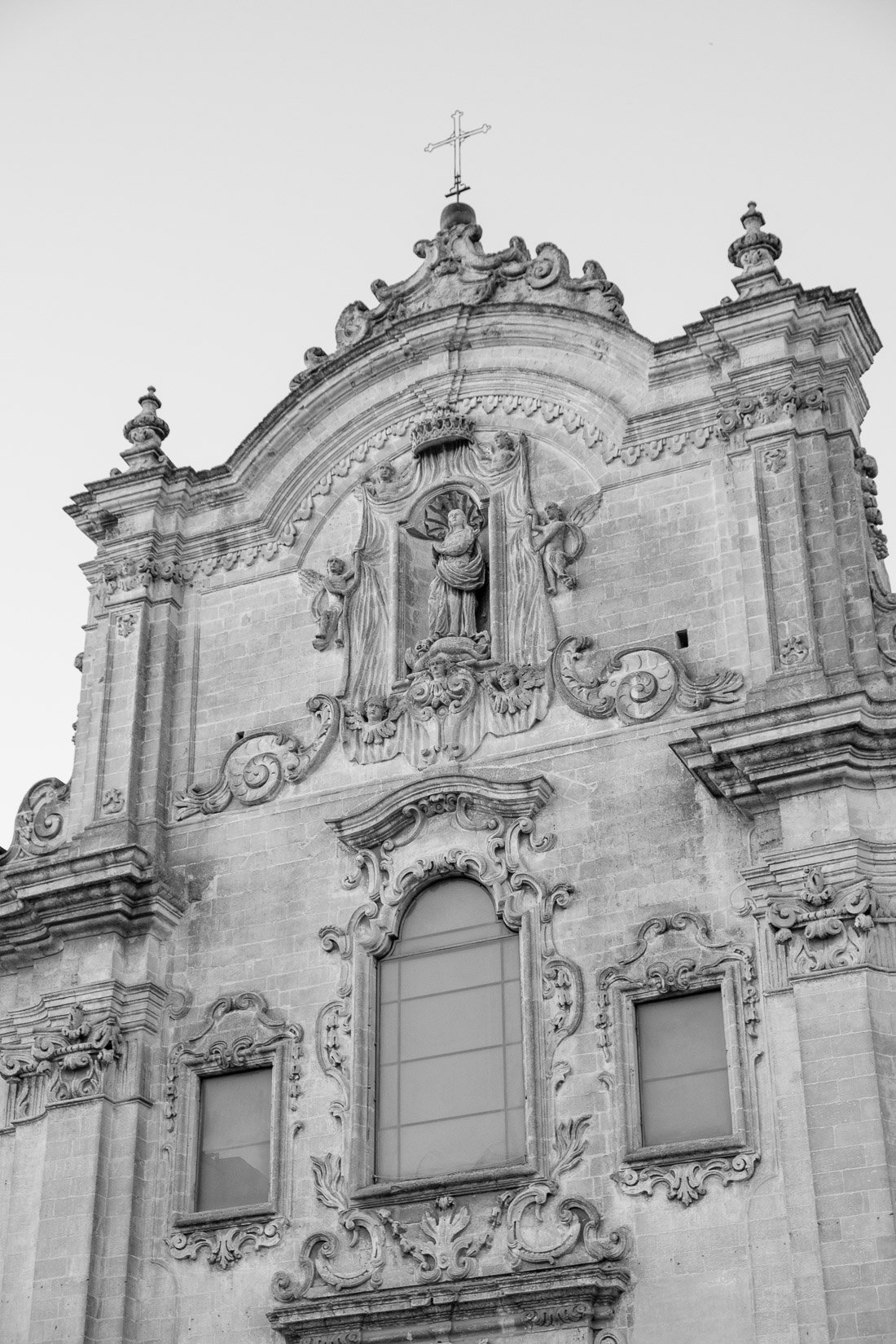 Black and white photo of an ornate, baroque-style church facade with sculptures of religious figures, elaborate carvings, decorative window frames, and a cross on top.