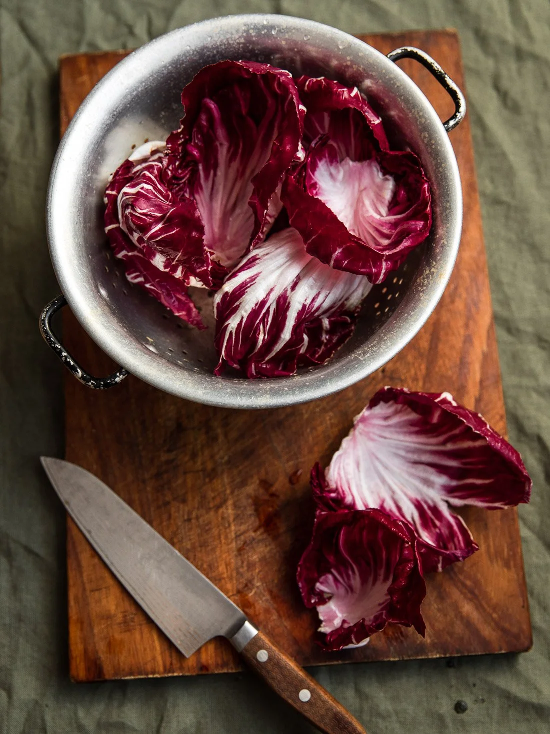 Radicchio leaves in a metal colander on a wooden cutting board with a kitchen knife beside them.