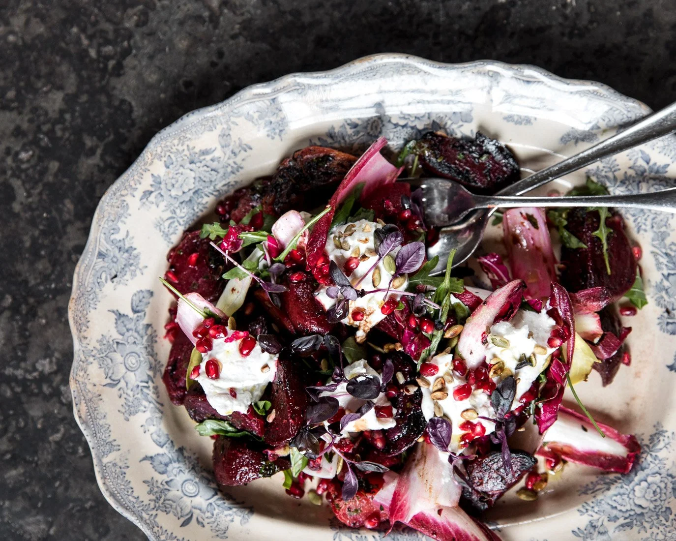 A decorative bowl containing a colorful salad with radicchio, microgreens, pomegranate seeds, walnuts, and dollops of white cheese, placed on a dark speckled surface.