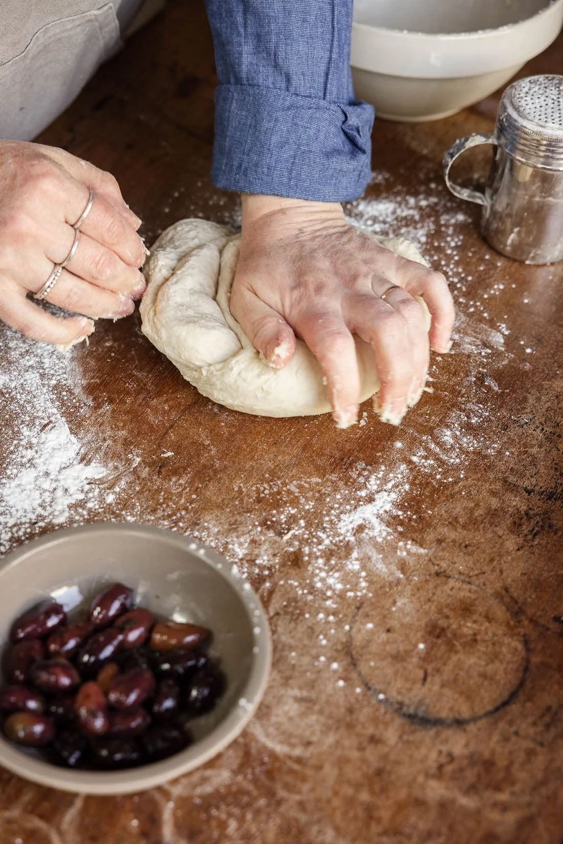 Person kneading dough on a floured wooden surface with a bowl of red beans nearby and a container of flour in the background.