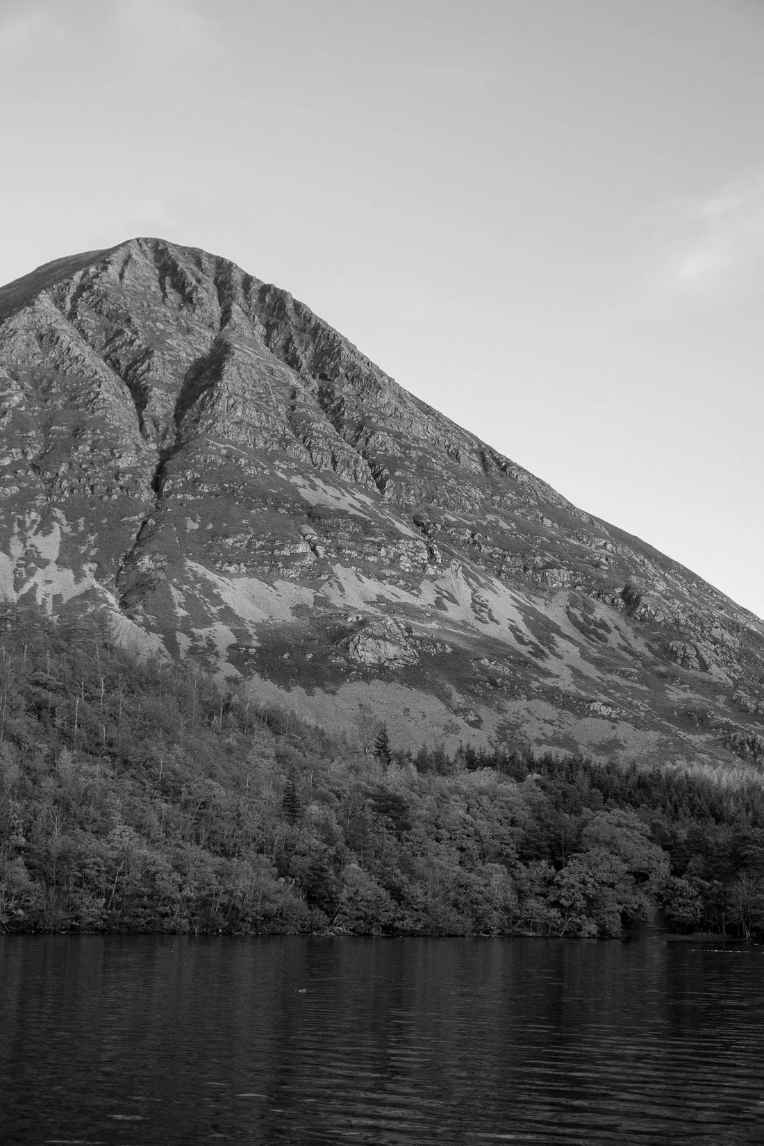 Black and white photograph of a mountain with a forested base and a body of water in the foreground.