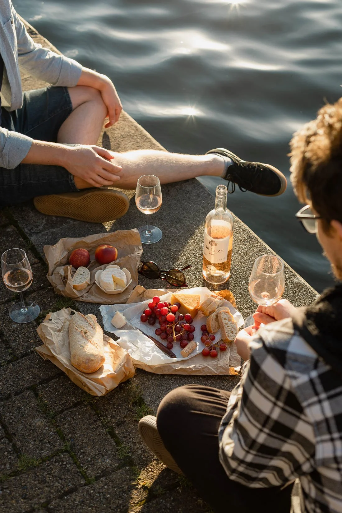 A picnic by the water with bread, cheese, grapes, apples, and wine, set on parchment paper on a paved surface, with two people sitting nearby, one holding a wine glass.