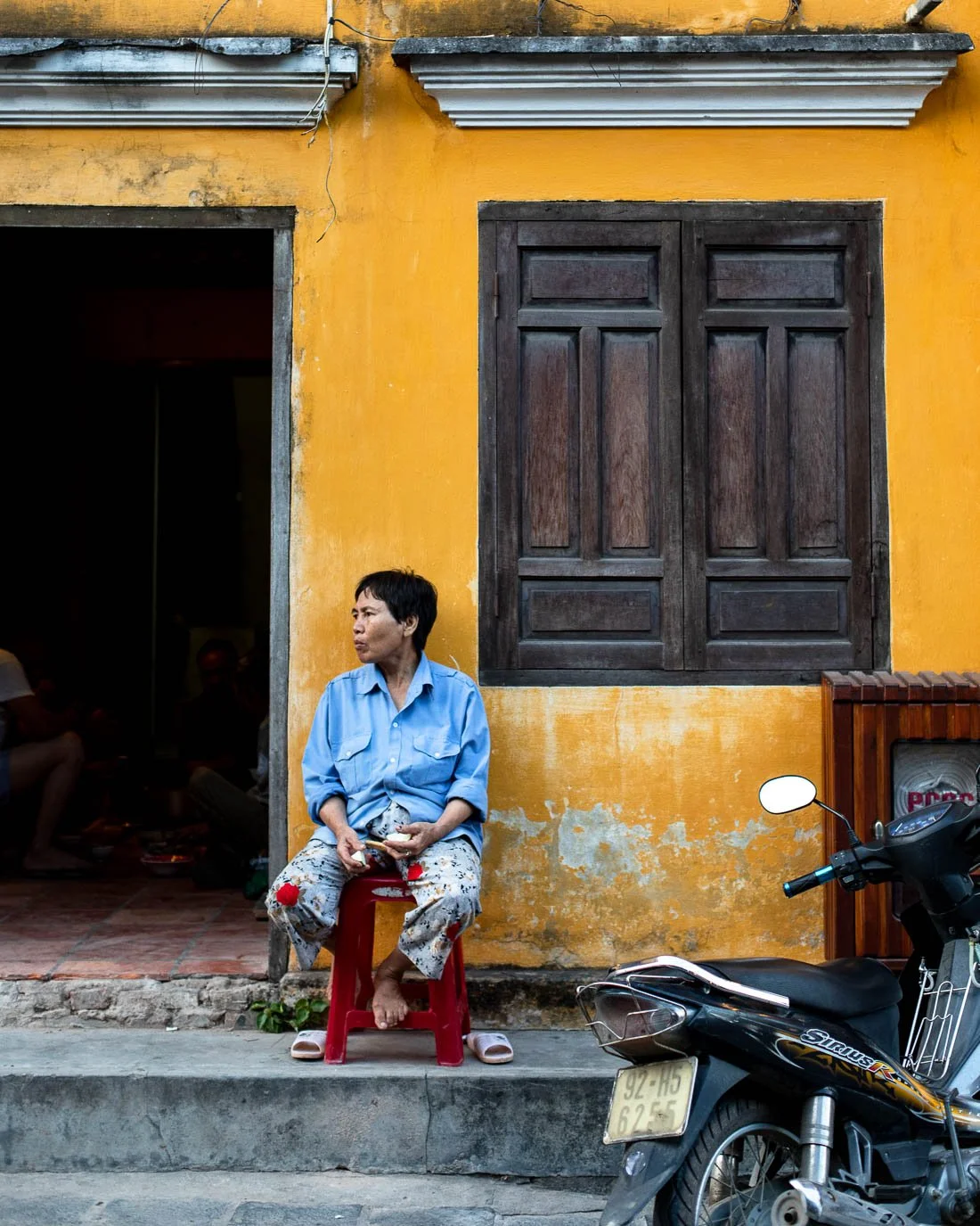 A woman sitting on a red plastic stool outside a yellow building with a large wooden window. She is wearing a blue shirt and colorful pants, holding what looks like a phone or small object. A motorcycle is parked nearby on the street.
